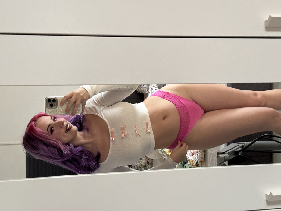 The image depicts an indoor mirror selfie taken in what appears to be a private room, potentially in the City of Zagreb, Croatia. A light-skinned individual, appearing female, is positioned facing forward, slightly angled left, smiling while capturing the image with a light-colored smartphone held in her right hand. Her hair is styled with distinct shades of purple and pink, and she wears dark lipstick. She is dressed in a white, long-sleeved, ribbed cropped top adorned with four small pink bows vertically aligned down the front. Her lower attire consists of bright pink briefs with lace detailing along the upper edge. Her left hand is positioned near the upper thigh of her left leg, slightly adjusting the garment. In the reflection behind the individual, a bed with bedding featuring a green and white floral or leaf pattern is partially visible on the left side. To the right and beneath her, dark cables and a dark object suggest the presence of electronic equipment or furniture. The room's background features plain, light-colored, possibly white, walls or wardrobe panels, contributing to a bright, minimalist environment.