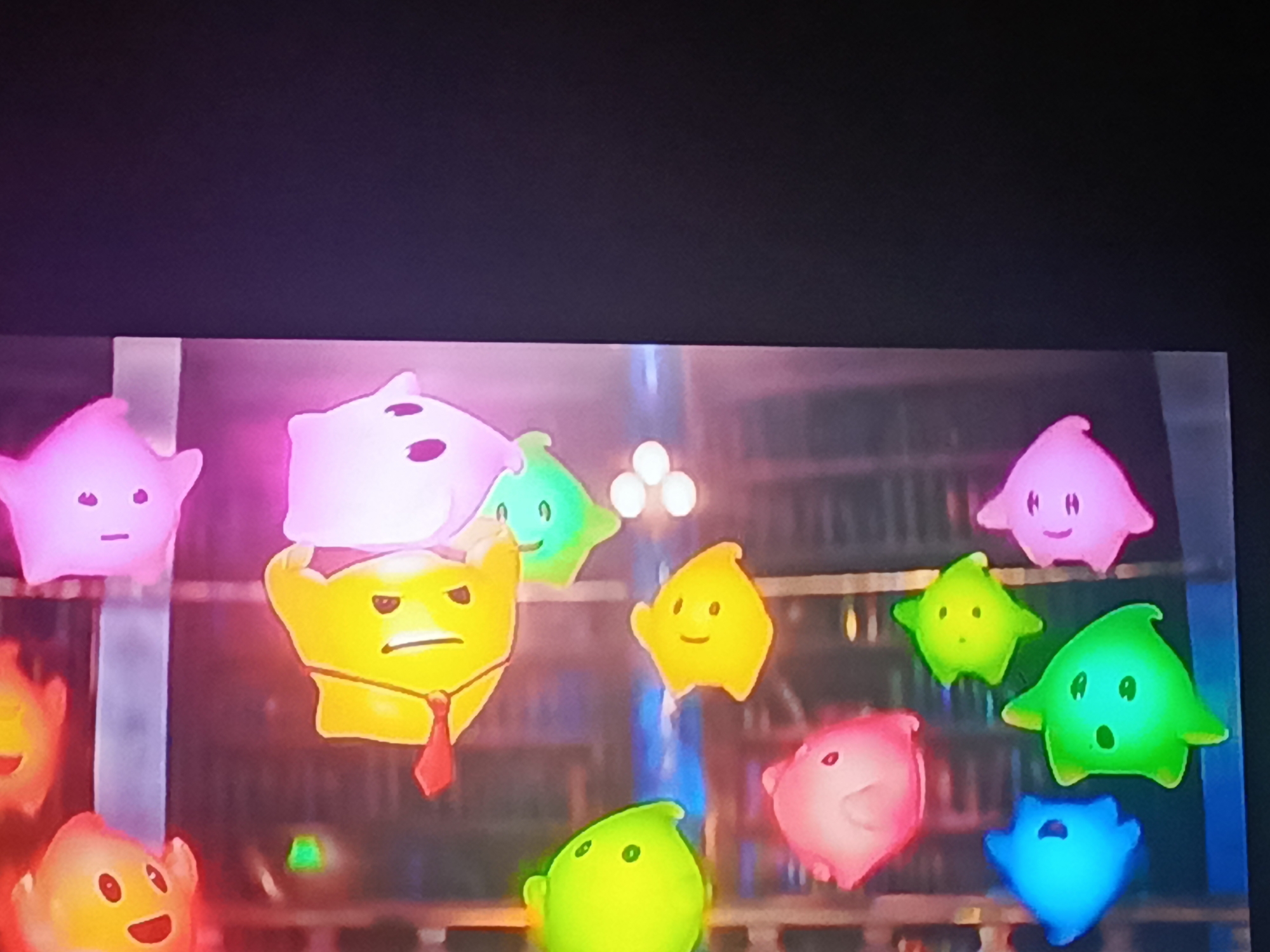 The image shows a screen displaying a scene with several brightly colored, cartoonish star-shaped figures. The figures are pink, yellow, green, and blue, each with different facial expressions. One yellow figure wears a red tie. The background appears to be a bookshelf or library interior with blurred details. The setting of Juan Griego, Venezuela, is not evident in the image.