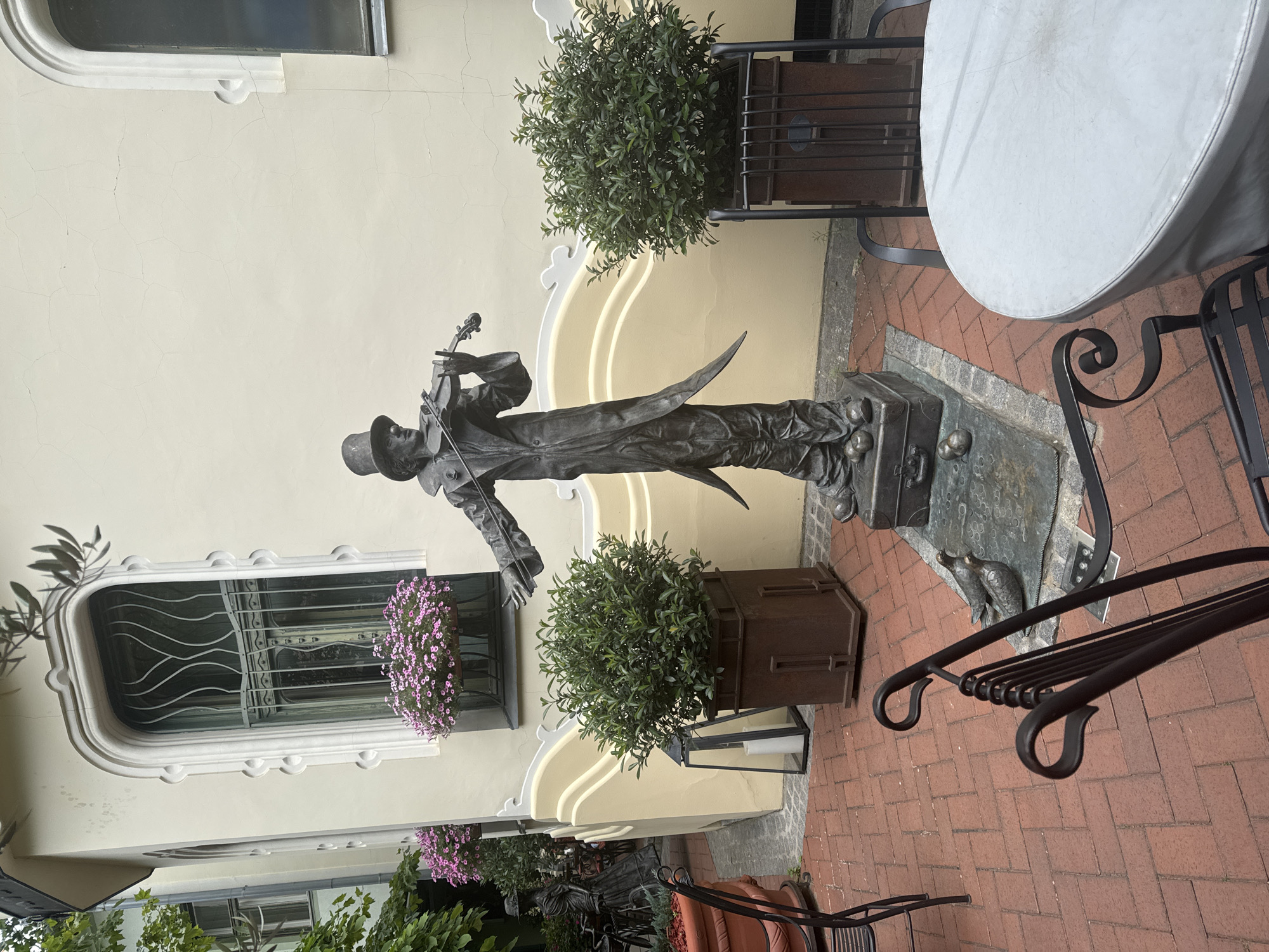 A bronze statue of a man playing a violin stands in a courtyard. The man wears a top hat and a long coat, and his stance suggests he is mid-performance. At his feet are several balls and a suitcase. Two bronze pigeons are positioned on a bronze plaque near the statue. The courtyard is paved with red brick and features potted green bushes, iron chairs, and a round table covered with a light-colored cloth. In the background, a cream-colored building with a distinct arched window adorned with flowering plants is visible. The architecture suggests a European style. This statue is located in Subotica, Serbia, and is known as the "Violinist" or "Boy with the Violin."