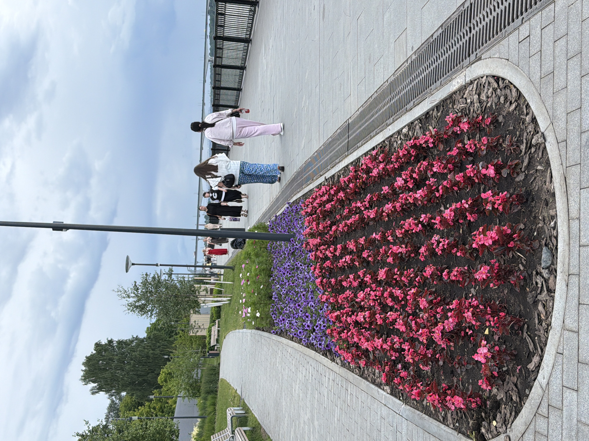 A group of people are walking along a paved walkway next to a flower bed. The flower bed is filled with bright pink and purple flowers and mulched with dark brown bark. In the background, there is a metal fence and a body of water. The sky is cloudy and the weather appears overcast. Some trees and lampposts are also visible in the scene. One person is wearing a pink jacket and pants, another a white jacket and a blue and white patterned skirt, and others are dressed in dark clothing. The people appear to be casually strolling along the walkway, possibly in a park or along a waterfront promenade in Perm, Russia.