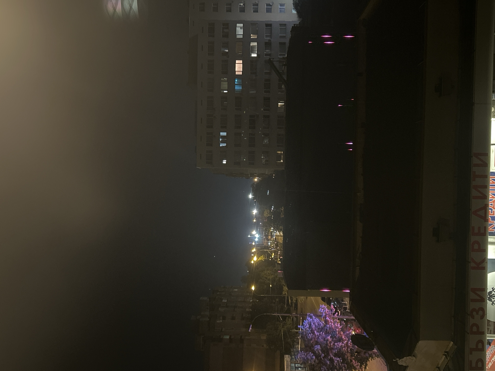 The image presents an urban night scene characterized by a widespread haze or fog. The location is the city of Sofia, Bulgaria, as confirmed by the prominent Cyrillic commercial sign "БЪРЗИ КРЕДИТИ" (Fast Credits) visible at the bottom of the frame, accompanied by partial text "CAMO CРЕЩУ..." (Only against...).
In the immediate foreground, dark, flat rooftops of lower-level buildings occupy the bottom half of the image. A satellite dish is mounted on the left-most rooftop. Below this rooftop and beside the street, a tree is distinctly illuminated with a purple light. Several thin, vertical light streaks, colored pink or magenta, are scattered across the dark rooftops in the mid-right foreground, possibly emanating from ground-level light sources or reflections.
The middle ground reveals an urban street or boulevard, lined on both sides by regularly spaced yellow-orange streetlights, which extend into the far distance. Distant vehicle lights are visible, indicating active traffic flow.
In the background, two large multi-story buildings dominate the horizon. The building on the left is dark in appearance, with a few windows showing internal illumination. The building on the right is a lighter-colored, extensively fenestrated structure, likely residential or mixed-use, with numerous windows, several of which are illuminated with distinct white, blue, and yellow light.
The sky is dark and appears significantly hazy or foggy, particularly towards the upper right, where a faint, diffuse glow from distant city lights is perceptible. No individuals are discernible within the frame. An optical artifact, possibly an internal reflection or flare from the camera's environment, is present in the upper right corner of the original image. The scene is static, captured at night, under artificial illumination and atmospheric conditions.