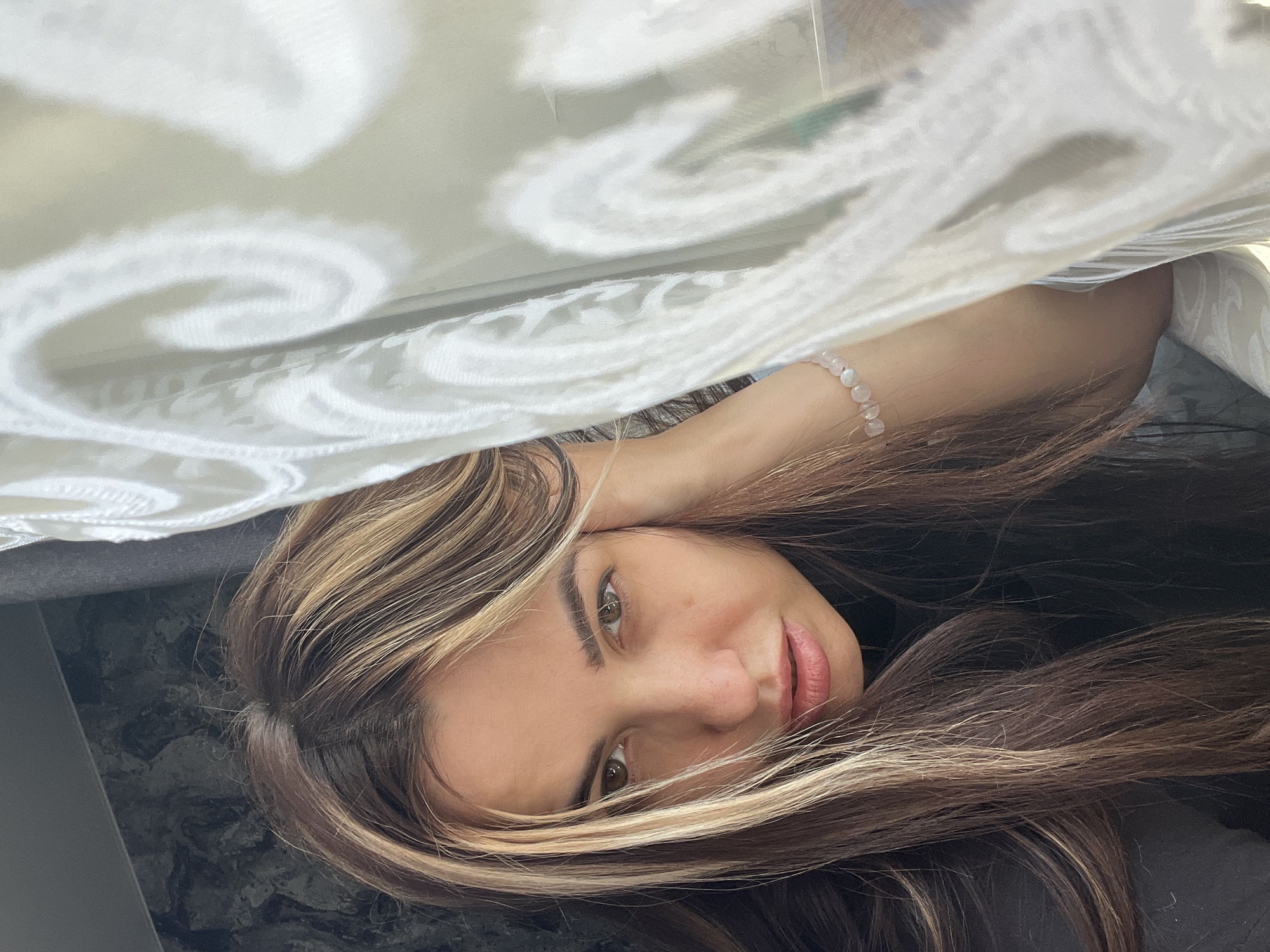 A young woman with light brown hair and blonde highlights is lying down. Her hair is partially covering her face. She is wearing a beaded bracelet on her left wrist. Above her is a white fabric with a patterned design. The location appears to be indoors. The city is unknown.