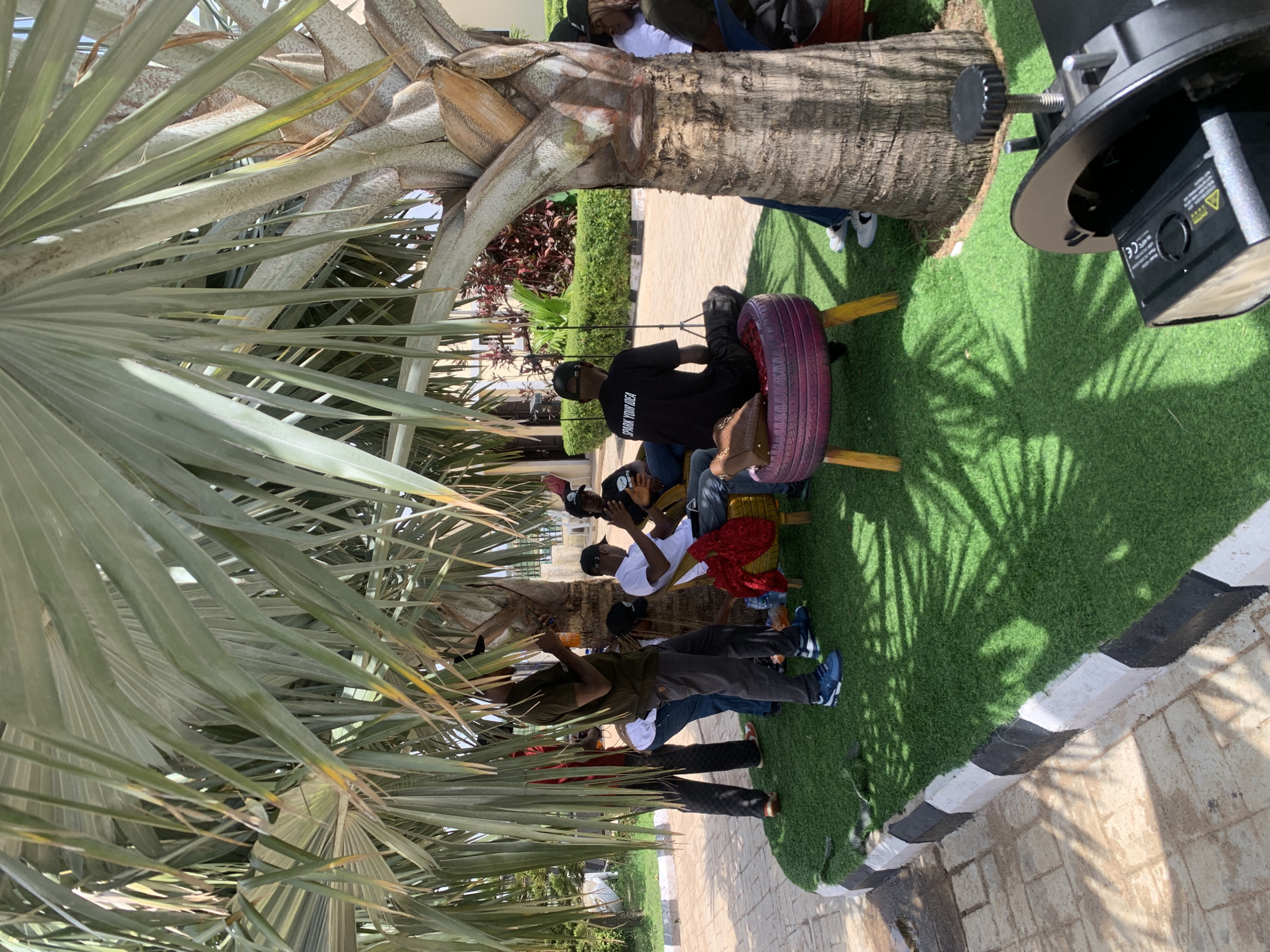 A group of young people are gathered outdoors under the shade of a large palm tree, enjoying what appears to be a casual social event or hangout. The scene is set on a patch of artificial turf, bordered by a paved walkway. The time of day is likely sunny, as indicated by the dappled shadows of the palm fronds on the ground.
Several individuals are visible, some sitting on a makeshift bench and others standing or sitting on the ground. One person is seated on a tire swing painted purple. Another individual in the foreground is wearing a dark shirt with the text "SPARK YOUR IDEA" visible on the back. Other individuals are wearing casual clothing like t-shirts and jeans.
The overall atmosphere seems relaxed and convivial. The presence of a camera or filming equipment in the upper right corner suggests that an event is being documented or that someone is engaged in photography or videography. The location context of Jos, Nigeria, places the scene in a specific geographic and cultural setting.
