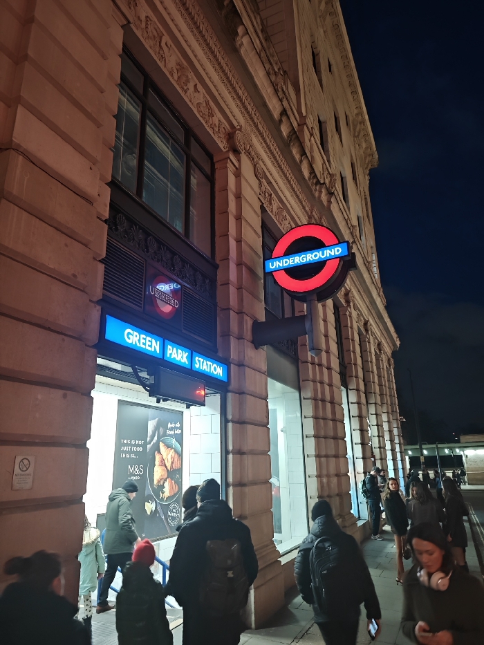 The image depicts a vibrant street scene at night, likely in an urban environment, centered around the entrance of a London Underground station. The time of day is clearly evening or night, indicated by the dark sky and the illuminated signs and building lights.
The prominent feature is a grand, multi-story building with classical architecture, characterized by light-colored stone, ornate cornices, decorative carvings along the frieze, and vertical pilasters or columns. The building's windows reflect the indoor lighting and the street scene.
At the entrance to the station, there are two distinct London Underground roundel signs. The most prominent one, on the right, is a large, circular red sign with a horizontal blue bar across its center, emblazoned with the word "UNDERGROUND" in white letters. A smaller, similar roundel sign is visible above the station entrance, reflecting on a glass surface, appearing somewhat distorted. Below this smaller roundel, a bright blue rectangular sign clearly reads "GREEN PARK STATION" in white, capital letters.
The station entrance itself appears to be integrated with a storefront, possibly a Marks & Spencer Food store, given the large poster visible through the glass window. This poster features text that reads "THIS IS NOT JUST FOOD THIS IS..." followed by "M&S FOOD" and a smaller "BAKERY" label. The poster displays an image of baked goods.
Numerous people are visible on the sidewalk in front of the station and the adjacent storefront. They are dressed in winter attire, including jackets, hats, and scarves, suggesting cool weather. Some individuals are walking past, while others appear to be entering or exiting the station/store. One person on the far right has headphones around their neck. The overall activity suggests a typical busy evening scene in a metropolitan area.
The sidewalk is paved, and further down the street on the right, streetlights are visible, illuminating more people and buildings in the distance. The atmosphere is one of urban movement and activity after dark, with the station and storefront acting as focal points of light and gathering. A faint circular sign with a crossed-out cigarette, indicating "no smoking," is visible on the wall to the left of the station entrance.