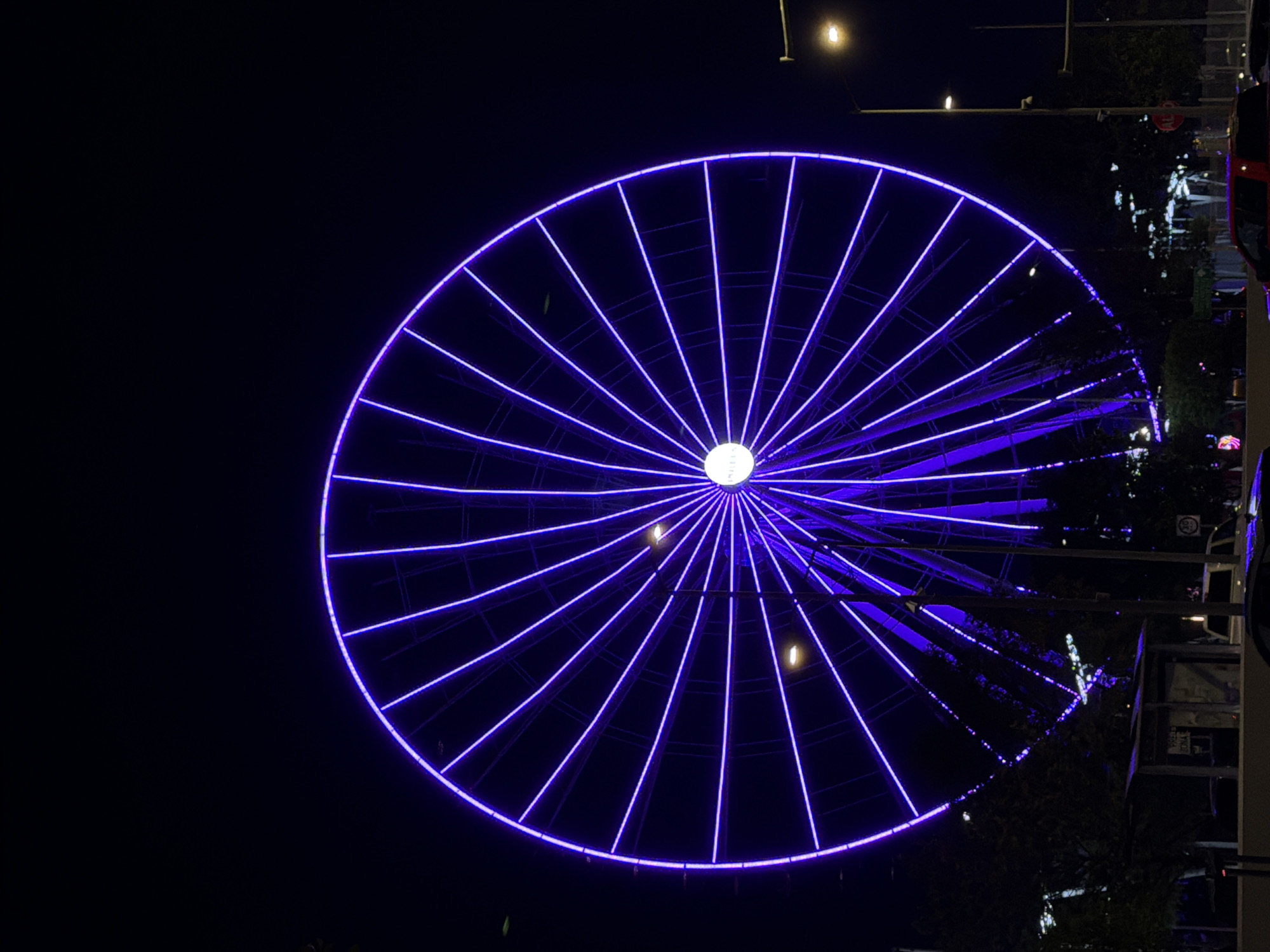 The image depicts La Estrella de Puebla (The Star of Puebla), a prominent Ferris wheel in the Zona Angelópolis of Puebla, Mexico, at night. The Ferris wheel dominates the frame, illuminated by numerous bright violet LED lights outlining its circular rim and radial spokes. Its central hub emits a distinct white light.
The sky is dark and clear. In the lower portion of the image, the tops of dark, silhouetted trees are visible, obscuring the base of the Ferris wheel. To the left, a bus stop shelter with partially visible text "PRELL ZONA" stands. Below and to the right of the bus stop, a red pickup truck is partially visible on what appears to be a paved road. Further right, a white SUV or van is parked or driving behind the bus stop. Multiple streetlights on tall poles are present, casting a warm yellow-orange glow, particularly on the right side of the image. A red "ALTO" (Stop) sign is mounted on a pole to the right of the bus stop structure. No individuals are discernible in the scene.