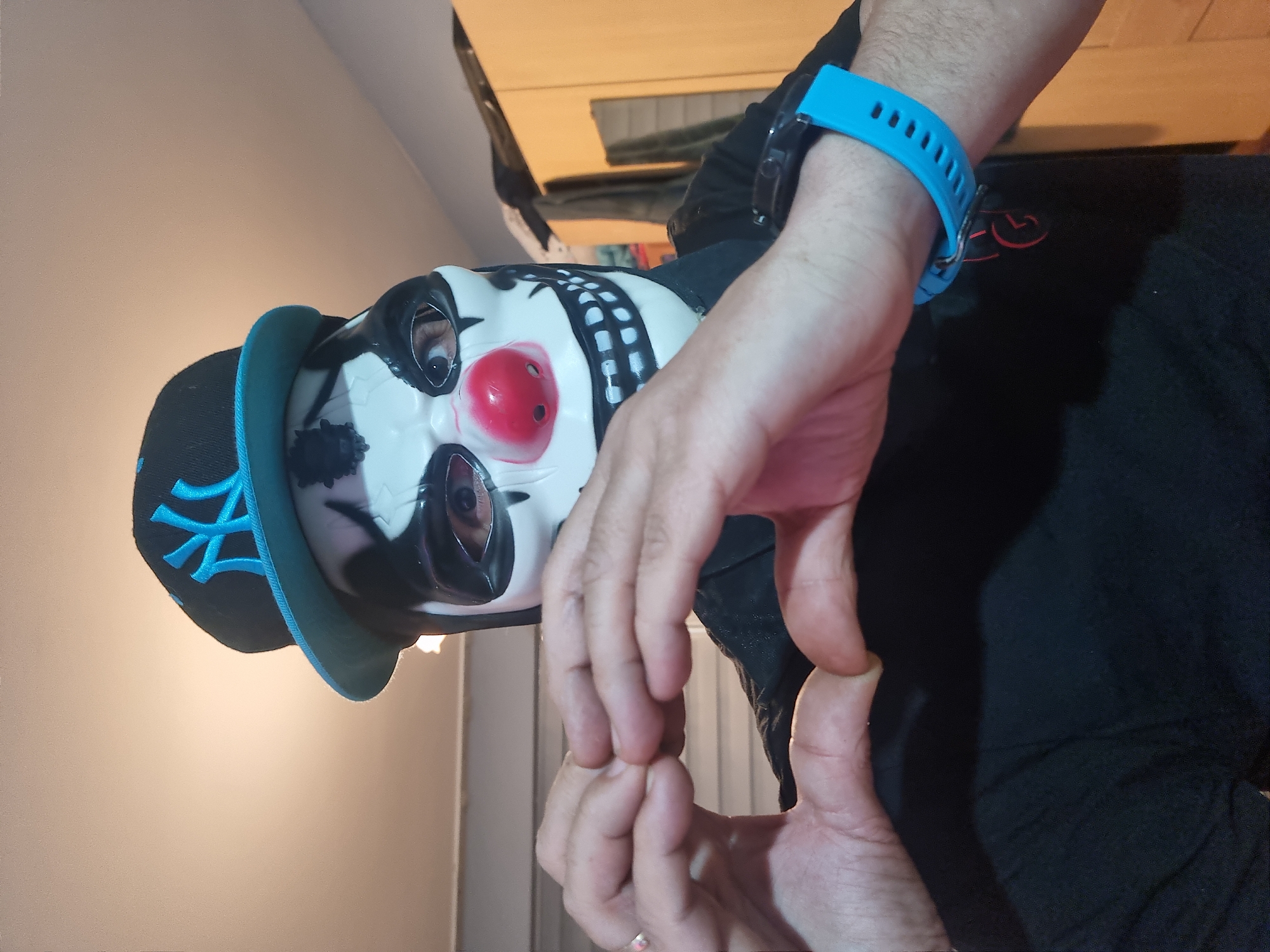 A person is wearing a clown mask and a black baseball cap with a blue logo and brim. They are also wearing a blue smartwatch. The person is wearing a black long-sleeved shirt. The background is a light-colored wall and a wooden cabinet. The location is unknown.