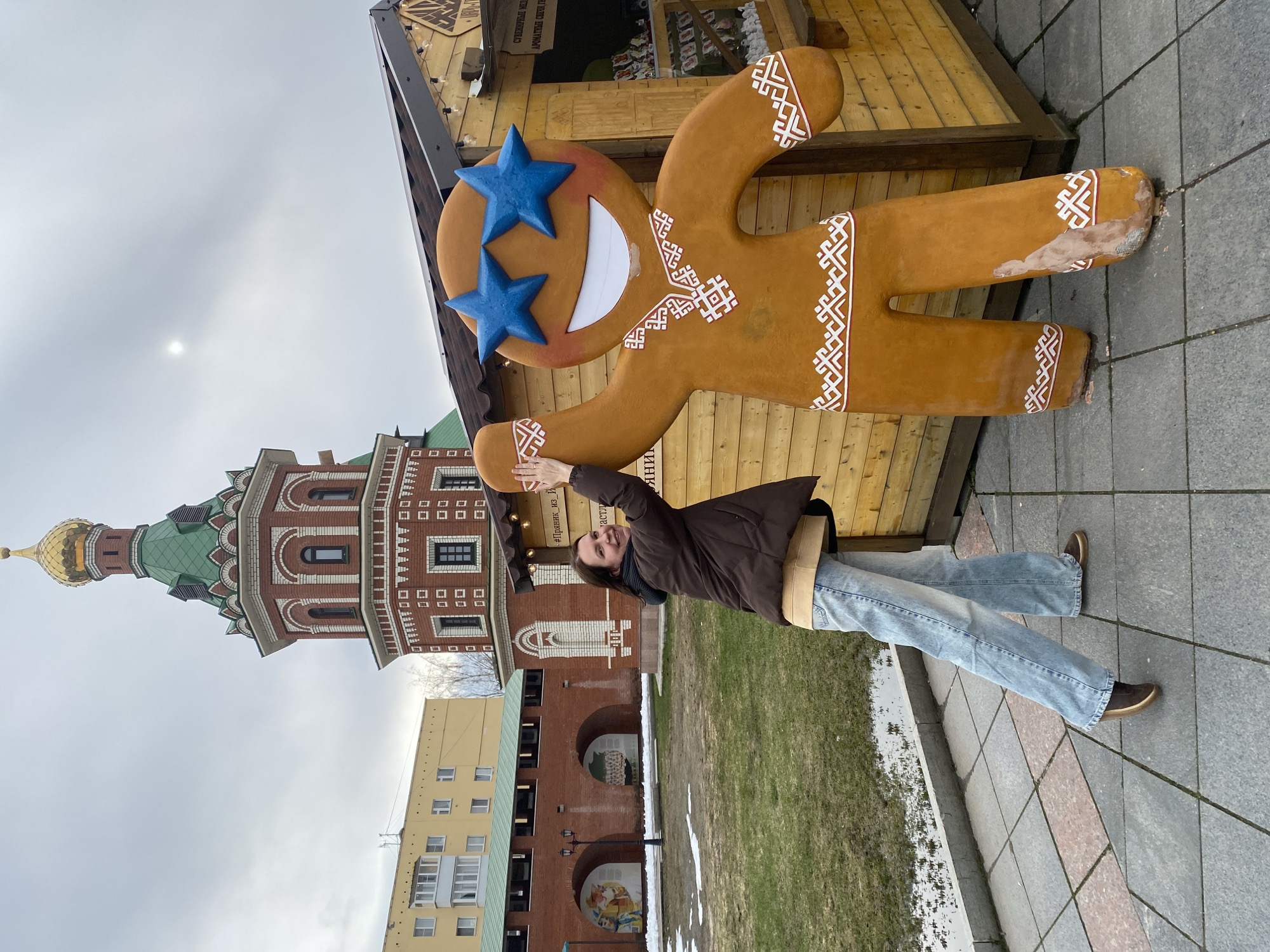 The image shows a young woman posing with a large gingerbread man cutout in Yoshkar-Ola, Russia. The gingerbread man has blue stars for eyes and is leaning against a wooden structure. The woman is standing to the left of the gingerbread man, with her arm raised towards it.
In the background, there's a distinctive red brick building with a green roof and a golden dome, a characteristic architectural style found in Yoshkar-Ola. A yellow building is also visible. The ground is paved with gray stones. The weather appears overcast. The image is rotated sideways. There is a sign partially visible on the wooden structure next to the gingerbread man.