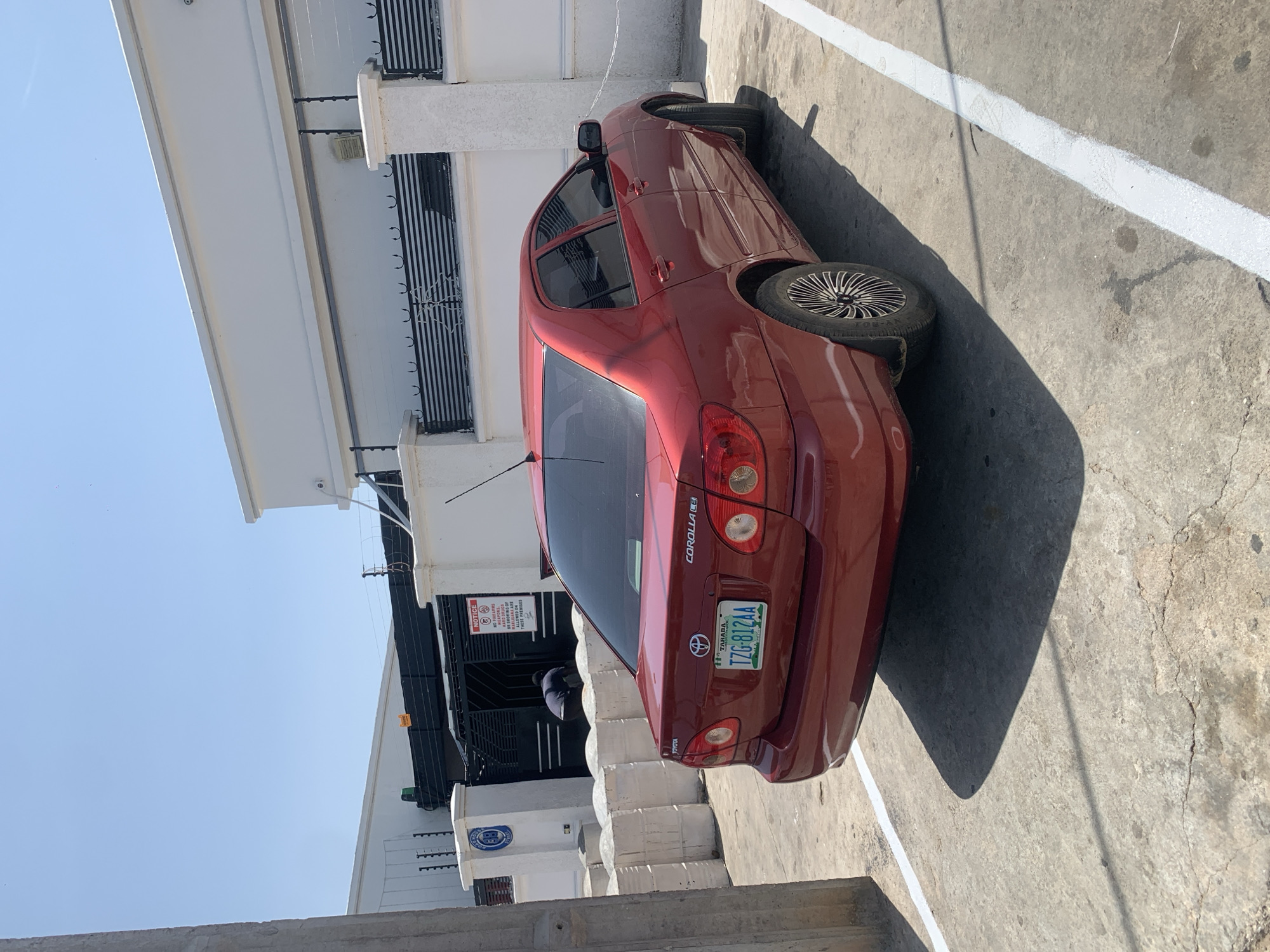 A red Toyota Corolla LE is parked on a concrete surface next to a white building with black security bars. The car's license plate reads "TZG-812AA" and indicates "TARABA." A "NOTICE" sign is visible on the building, stating "NO FIREARMS OF SMOKING OF MARIJUANA ARE ALLOWED ON THESE PREMISES." A person in dark clothing is bent over in the doorway of the building. The scene appears to be outdoors in daylight, with a clear blue sky. The image is oriented with the top of the frame on the left, so the car and building are positioned sideways relative to a typical viewing orientation. The concrete ground is marked with a white line, suggesting a parking space. The overall atmosphere is mundane and appears to be in a secured or private area.