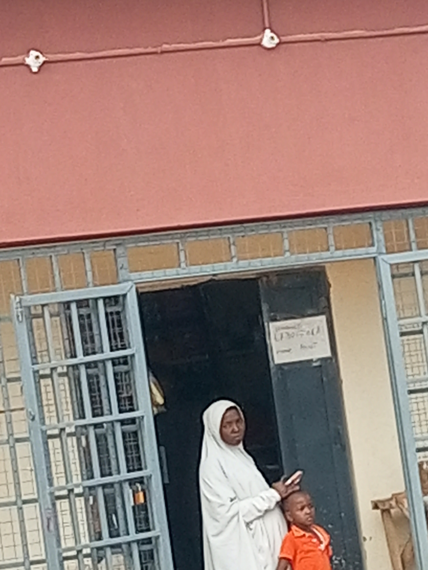 A woman wearing a white hijab and robe stands next to a young boy in an orange shirt. They are standing in front of a building with a reddish-brown facade and a dark doorway. The woman appears to be holding a phone. A sign with text is affixed to the doorframe to the right of the doorway. A metal grate covers a window to the left of the doorway.