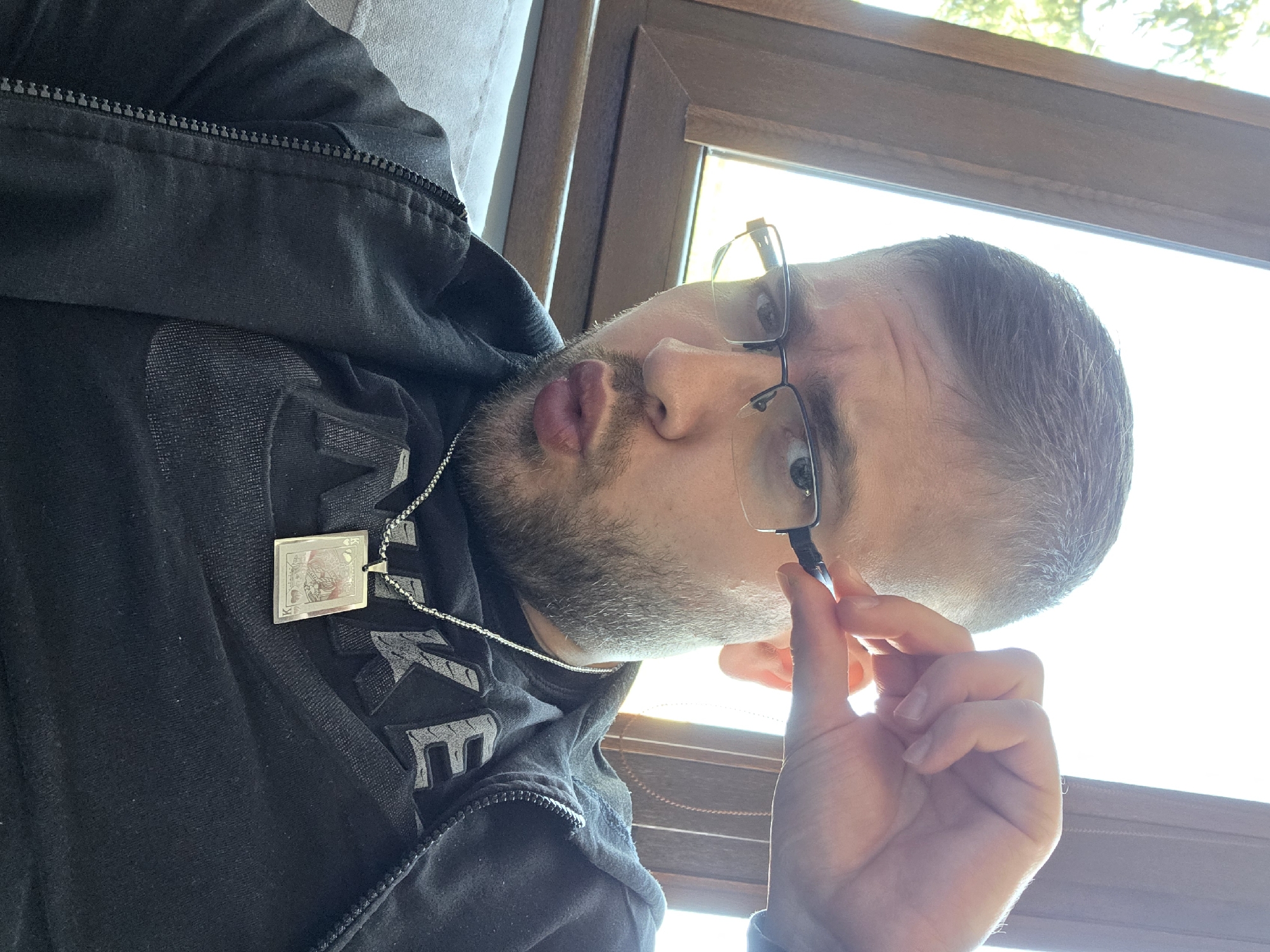 A man with short, light brown hair and a trimmed beard is looking at the camera. He's wearing glasses and a black zip-up hoodie over a black t-shirt with the word "NIKE" in gray letters. Around his neck is a silver chain with a square pendant resembling a playing card. He's holding the temple of his glasses with his right hand. The background shows a window with light streaming through, possibly suggesting daylight. The image was taken in Yagodovo, Bulgaria.