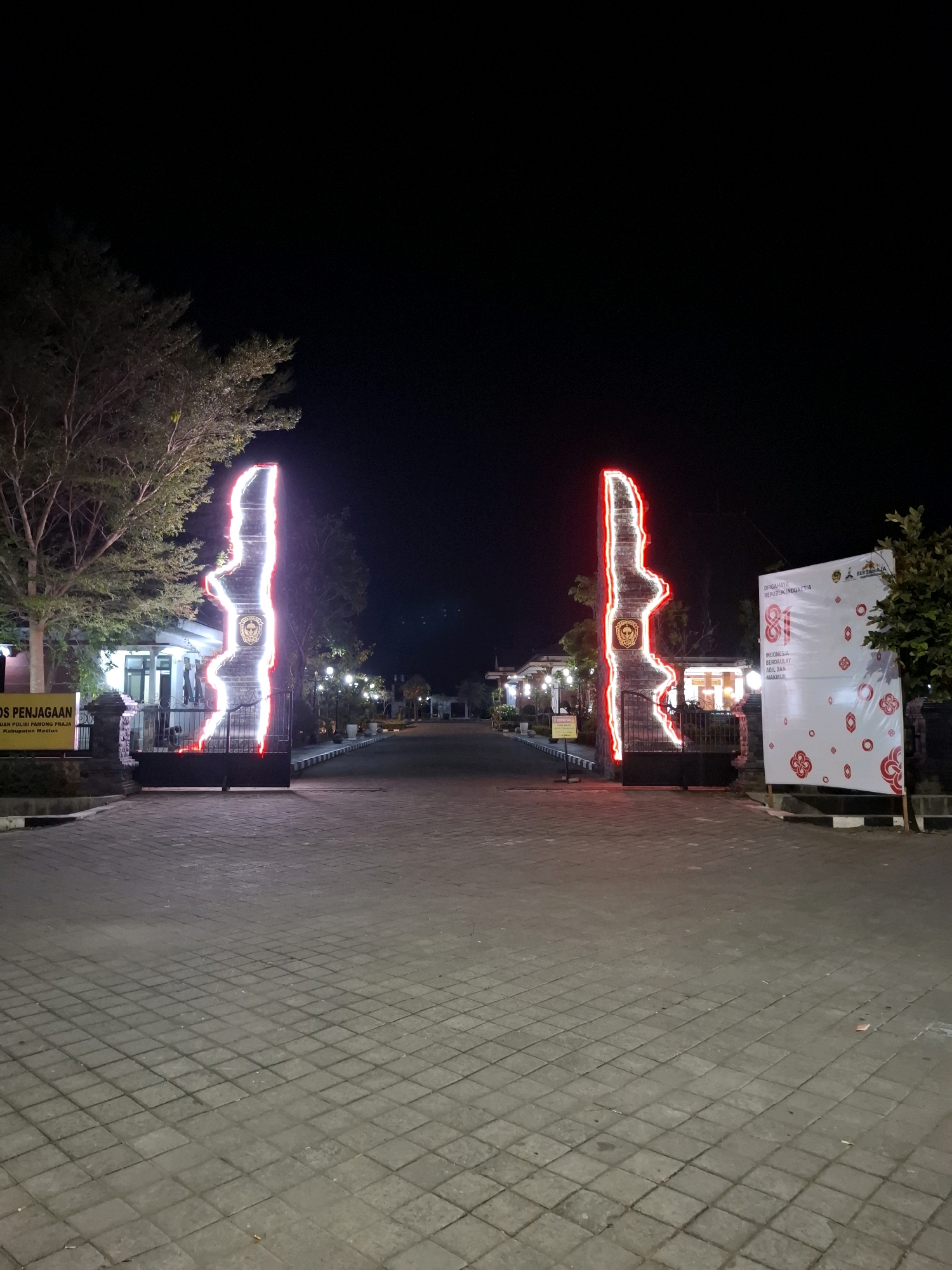 The image shows an entrance gate at night in Pandean, Indonesia. Two tall, slender structures, likely pillars, are adorned with bright red and white neon lights outlining their irregular shapes. The left pillar has a circular emblem at its center. The gate itself is a dark metal structure. In front of the left pillar, a yellow sign with black text, which includes the words "POS PENJAGAAN," is visible. To the right of the right pillar, a large white banner is displayed, featuring red floral motifs and text in Indonesian, including "DIRGAHAYU REPUBLIK INDONESIA." The ground in front of the gate is paved with square tiles. Trees with sparse foliage are present on the left side, and lights illuminate the background, suggesting buildings and pathways. The sky is dark, indicating nighttime.