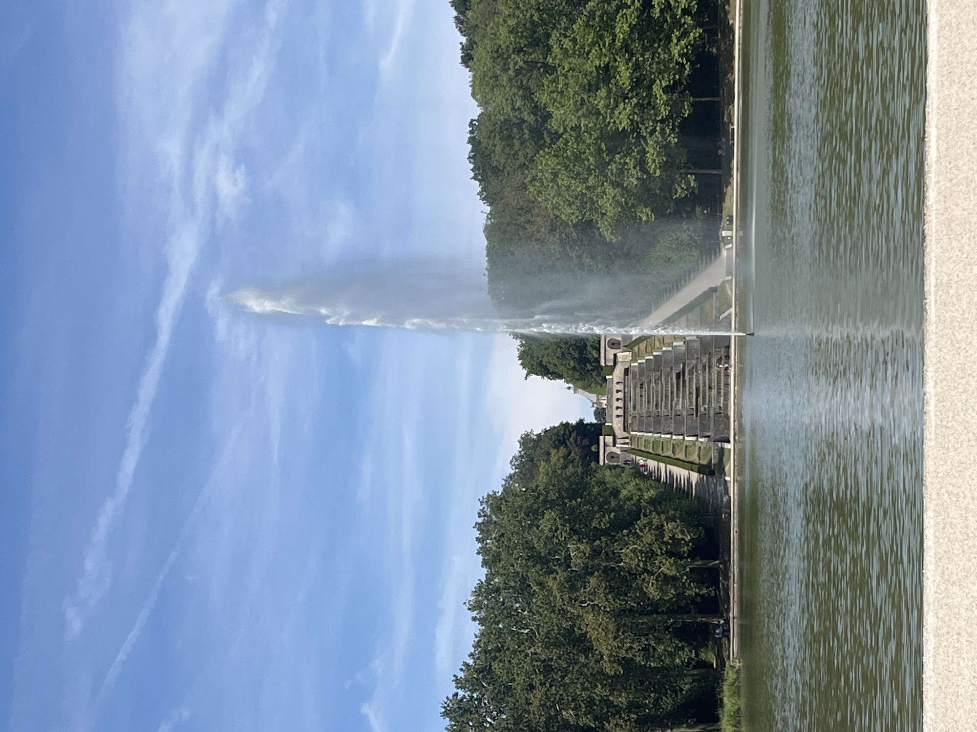 This is a full, eye-level, vertical outdoor shot of a large, tiered water fountain in the Parc de Sceaux, located in Antony, France. The fountain is the dominant subject, with a tall, powerful stream of water shooting upwards from the center of a pond in the foreground. The pond is calm with rippling surface. Behind the pond, a grand stone staircase cascades down from a higher elevation, with water flowing over its many steps. This staircase is flanked by lush green trees on both sides. The sky above is blue with scattered white clouds, some of which appear to be contrails. There are several people visible on the staircase and on benches near the pond, enjoying the scenery. The overall atmosphere is serene and picturesque, suggesting a beautiful summer day.
