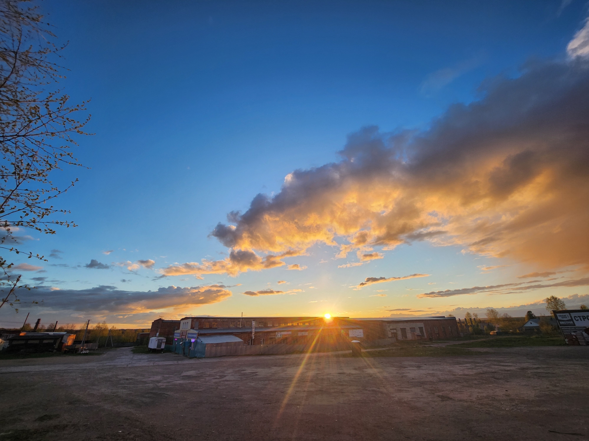 The image shows a sunset in what appears to be a rural or industrial area of Krasnogorskoye, Russia. The sky is a gradient of blue transitioning to orange and pink near the horizon, with clouds illuminated by the setting sun. In the foreground, there's an unpaved area, possibly dirt or gravel. A low, long building is silhouetted against the sunset. Adjacent to the building are some small structures, possibly sheds or outbuildings. A tree branch with budding leaves is visible in the upper left corner of the frame. The overall scene conveys a sense of quietude and fading light.