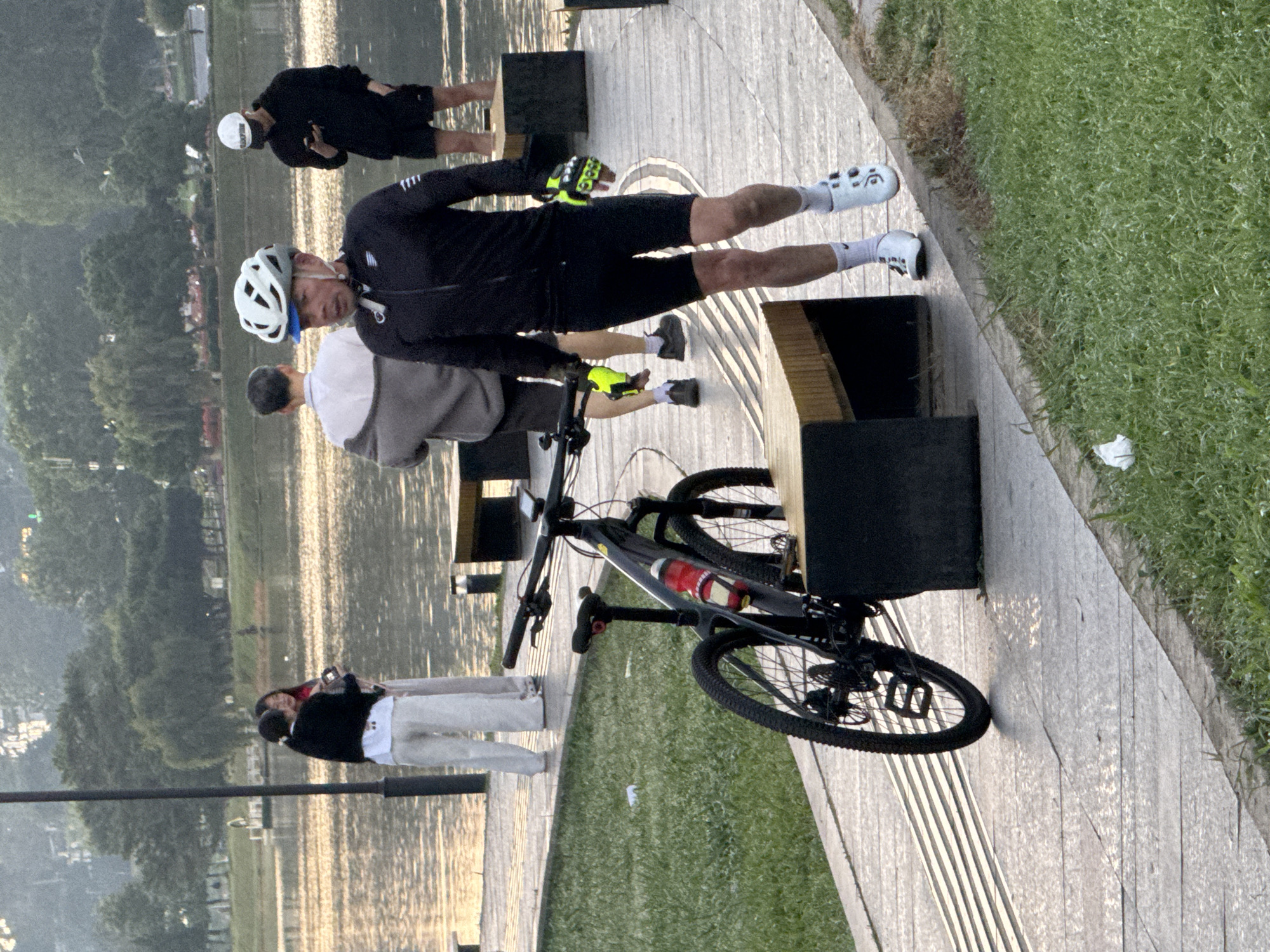 The image depicts Xuan Huong Lake in Da Lat, Vietnam, at sunrise. The golden light of the rising sun reflects off the water, creating a serene atmosphere. In the foreground, a man in cycling attire, including a helmet and gloves, walks towards the viewer, his bicycle parked nearby. His black cycling jacket and shorts are visible, along with white cycling shoes and socks.
Behind him, two individuals stand near the water's edge, their backs to the camera. The person on the left wears light-colored pants and a dark jacket over a white shirt, while the person on the right has on dark shorts and a black hoodie, holding a phone. Further in the background, another person in a grey jacket and dark shorts stands near the water, facing away from the camera. The scene is set on a paved walkway lined with benches and grass, with trees and hills forming the backdrop.