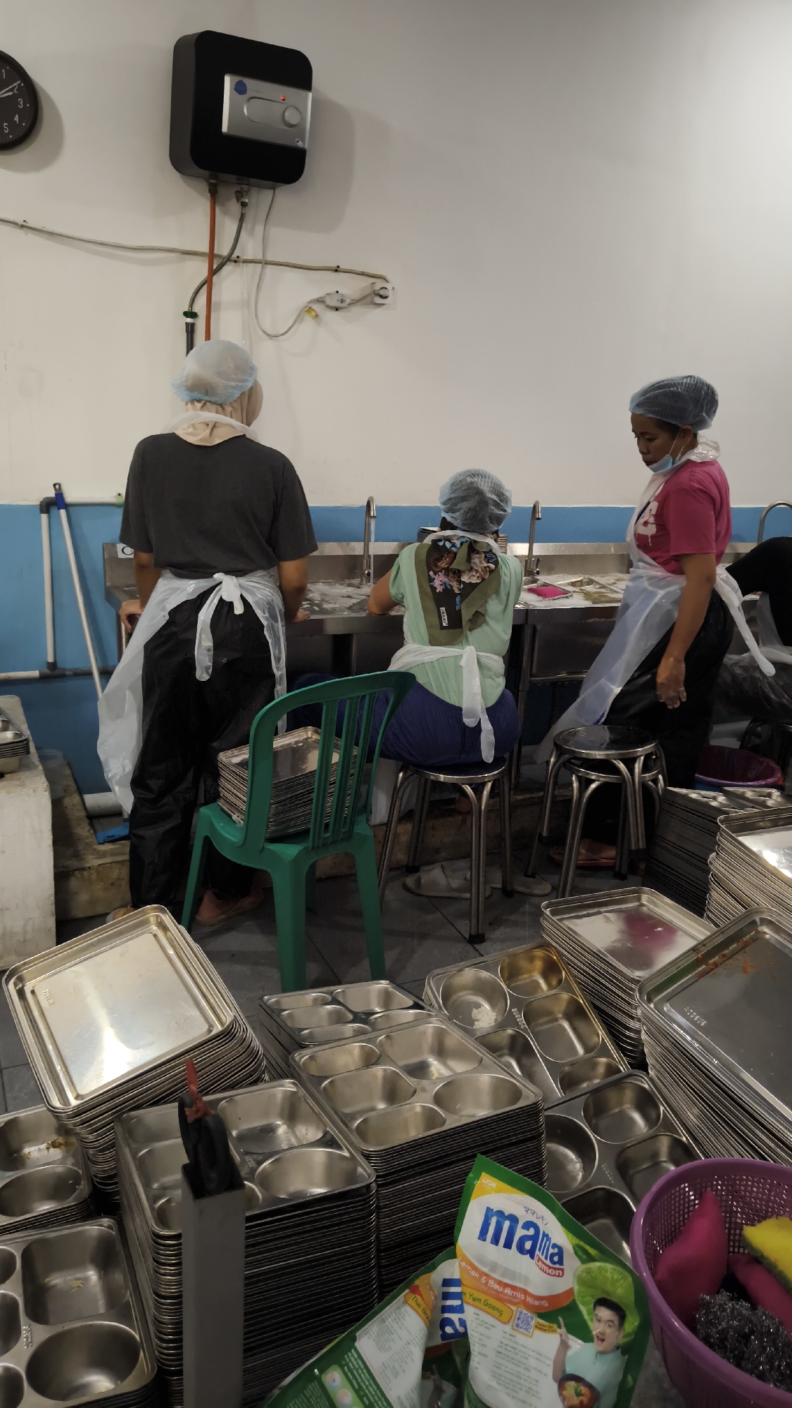 In Sambipondok, Indonesia, three individuals are observed in what appears to be a kitchen or food preparation area. The scene is indoors, with white walls and blue wainscoting along the lower portion. A prominent electric water heater is mounted on the upper section of the wall.
One person, wearing a dark t-shirt and a plastic apron over black pants, stands facing away from the camera, engaged in an activity at a stainless steel sink. They are also wearing a hairnet.
Another individual, seated on a stool and facing away from the camera, is wearing a light green shirt and a plastic apron, also with a hairnet. This person is working at a sink.
A third person, standing and facing the camera from the right, is wearing a pink shirt and a plastic apron. This individual is also wearing a hairnet and appears to be observing or interacting with the others.
The foreground of the image is dominated by stacks of metal trays, some of which are multi-compartment. A green plastic chair is positioned in front of the standing individual. A purple basket containing cleaning supplies, including a sponge and scrub brush, is visible to the right. Packaging for a product named "mama lemon" is also present in the lower right portion of the image. The overall impression is a functional food service environment with clear sanitation protocols being followed.