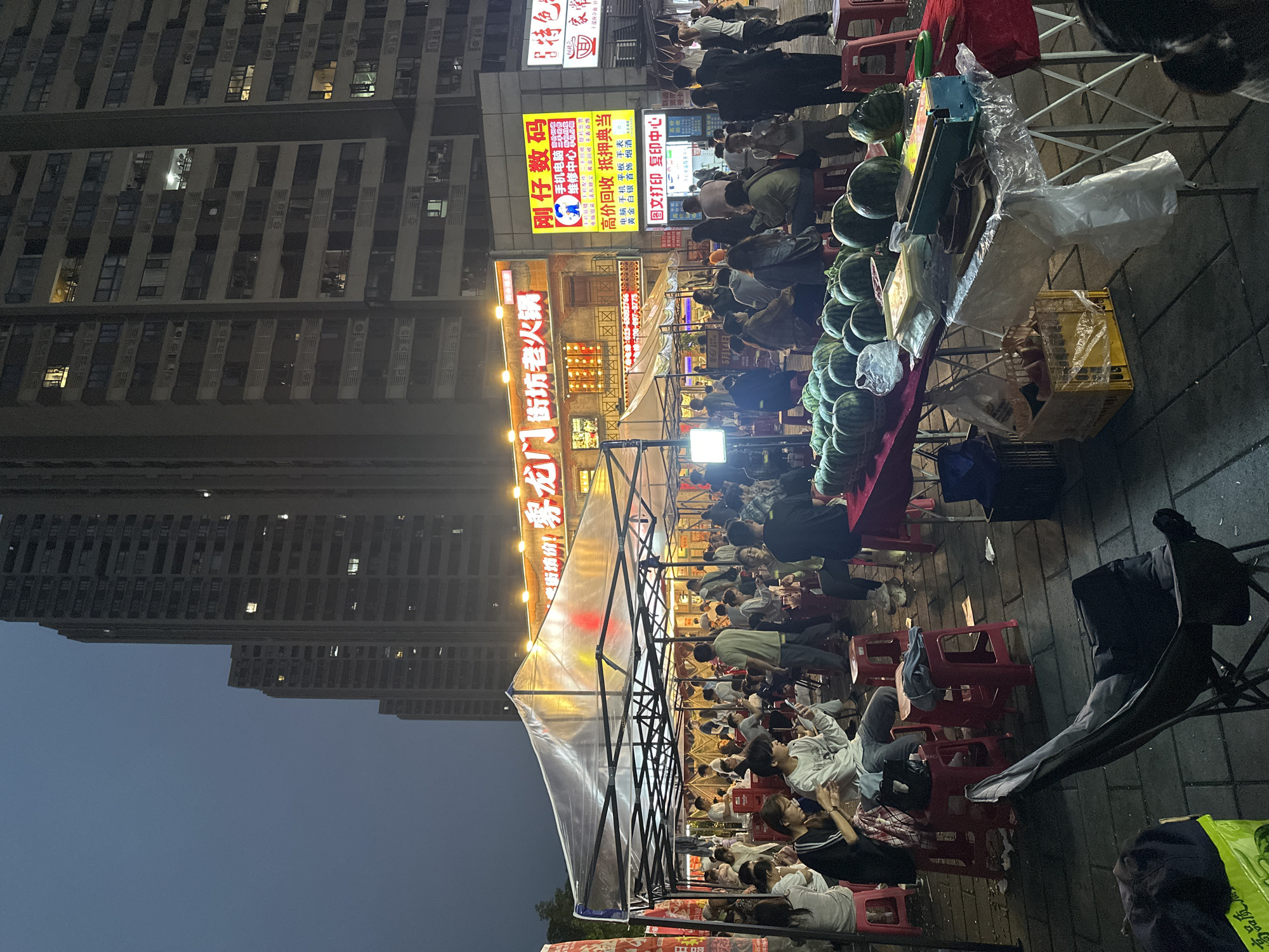 The image depicts an outdoor food market scene in Chongqing High-tech Zone, China, at night. Tall apartment buildings loom in the background under a dark sky. Several people are seated at tables under a tented structure, suggesting a restaurant or food stall setup. Red plastic stools are scattered around the tables. In the foreground, there's a table with a display of watermelons. Illuminated signs in Chinese characters are visible above the food stalls, with "龙门 街坊老火锅" being the most prominent. There are also signs for a repair shop and a printing service, along with more people in the distance. A few bags are on the sidewalk.