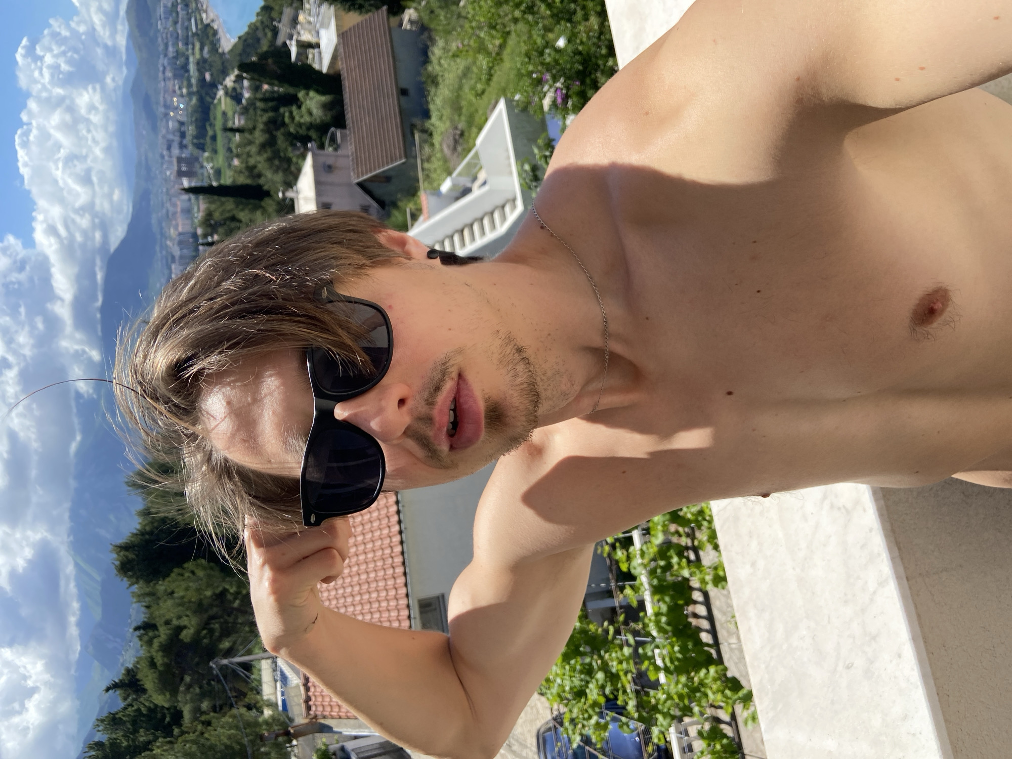 A medium close-up, high-angle shot depicts a shirtless man with light skin, brown hair, and sunglasses, appearing to pose for a selfie outdoors. He is positioned near a ledge or balcony, with an arm flexed, suggestive of showcasing his bicep. A silver chain is visible around his neck.
The background reveals a cityscape with buildings, trees, and distant mountains under a blue sky with white clouds. Specific details suggest the location is likely Šušanj, Montenegro: the architecture, terrain, and vegetation align with the typical characteristics of the coastal town. Orange terracotta tiled rooftops are visible among the greenery, further supporting this location.