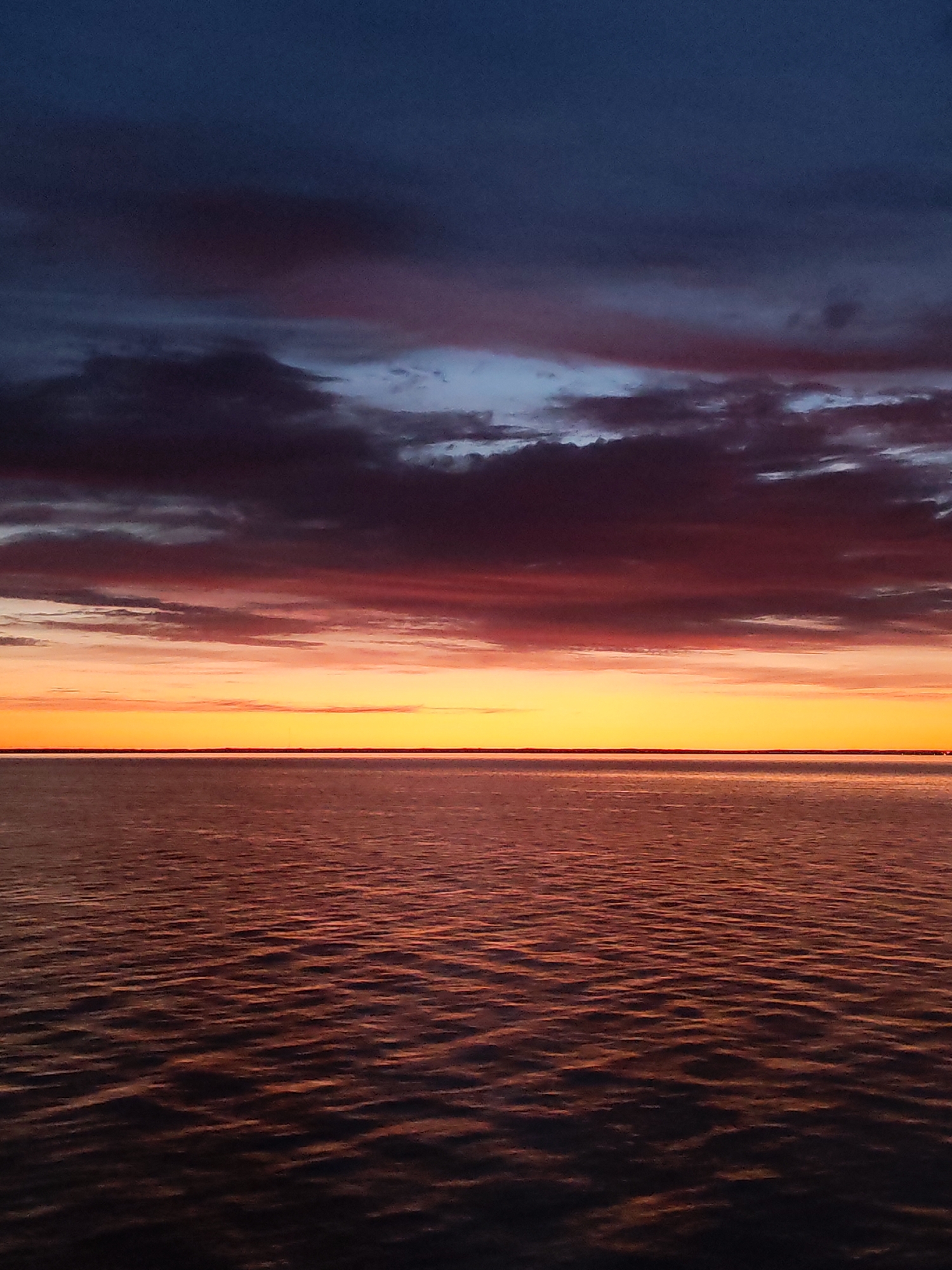 The image depicts a sunset over a body of water, likely a lake or the sea. The sky is a gradient of colors, transitioning from dark blue at the top to reddish-orange near the horizon. Clouds are present, exhibiting hues of purple, red, and gray. The water reflects the colors of the sunset, with ripples and waves creating a textured surface. The horizon line is sharp and distinct, separating the sky from the water. The location is specified as Unknown, Russia. No people, subjects, or objects are visible beyond the natural elements.