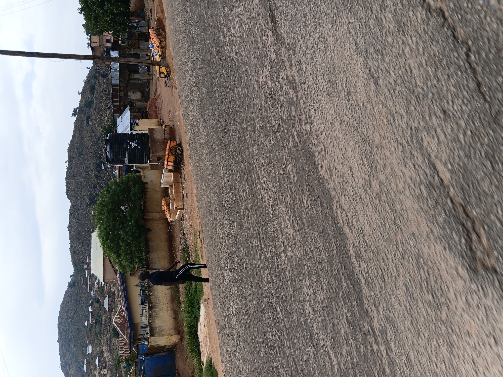 The image shows a road in Jos, Nigeria, with a person walking on the side. The road is gray and appears to be asphalt. On the left side of the road, there are buildings and a hillside with vegetation. One building has a water tank. There are a few carts. The sky is overcast. The person is wearing dark clothing with white stripes on the pants.