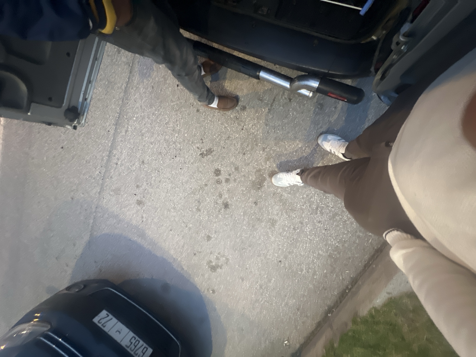 The image, taken from an overhead perspective in Martil ⵎⴰⵔْتِيل مرتيل, Morocco, shows three individuals near two parked vehicles on a concrete surface. One person wears brown loafers with white socks, grey pants, and a dark blue top with a yellow band on the sleeve. Another person wears white sneakers and brown pants, and is partially obscured by a person in white pants and a white top. A black exhaust pipe lies on the ground between the vehicles. The license plate on the car is visible, reading "6765 ⲩ 72." The background shows a strip of grass along the edge of the concrete.