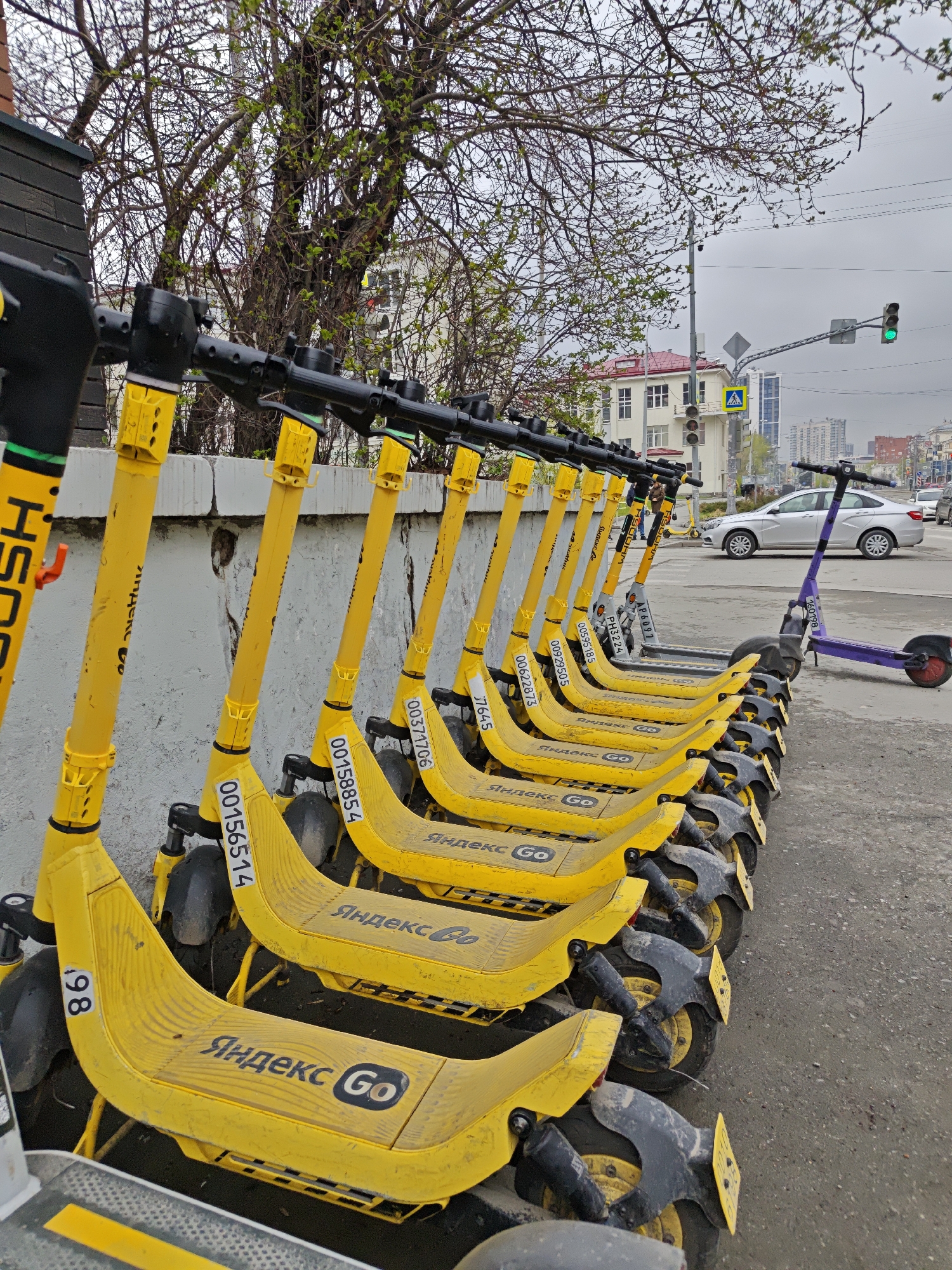 The image shows a row of yellow electric scooters parked on the sidewalk in Yekaterinburg, Russia. Each scooter has "Яндекс Go" written in Cyrillic on its base. The scooters are parked along a gray concrete wall. Beyond the scooters and the wall, there is a street with light-colored cars driving on it. In the background, there are buildings of varying heights, and a traffic light showing a green light. The sky is overcast. There is one purple scooter at the far end of the row.