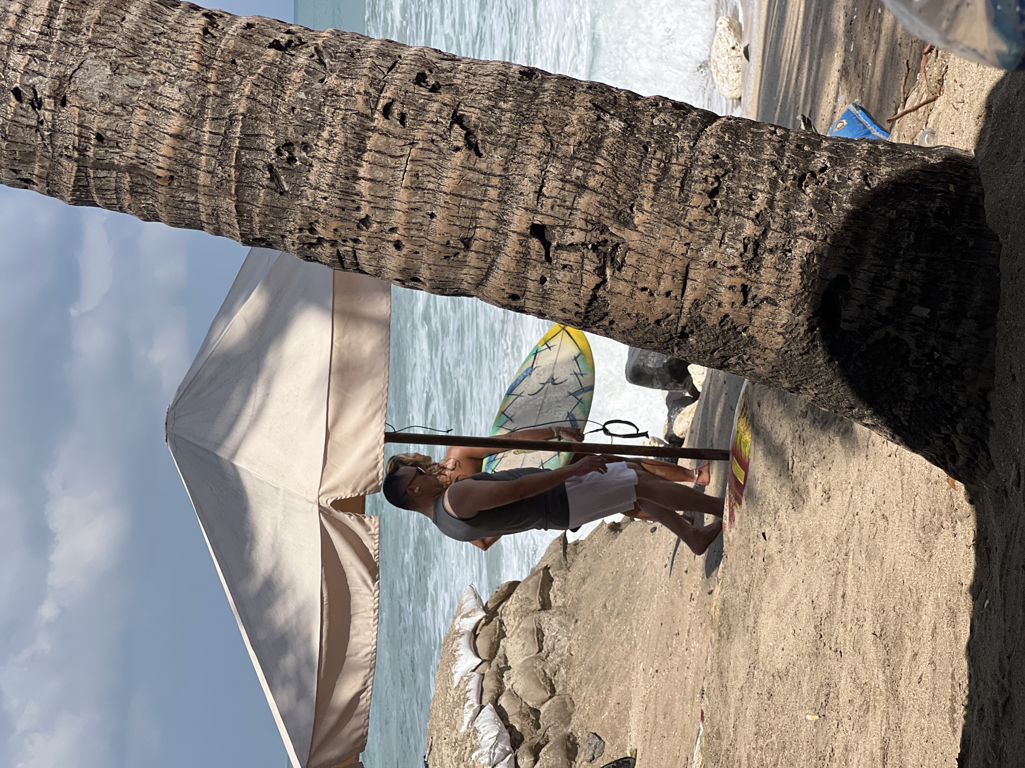 A man and a woman are standing on a sandy beach under a large, tan umbrella in Kuta, Indonesia. The man, wearing a dark sleeveless shirt and white shorts, is facing away from the camera. The woman, in the background, is holding a surfboard. The ocean with moderate waves is visible behind them. In the foreground, a large palm tree trunk is prominent, casting a shadow. Sandbags are stacked to the left, suggesting a potential sea defense. The sky is partly cloudy.