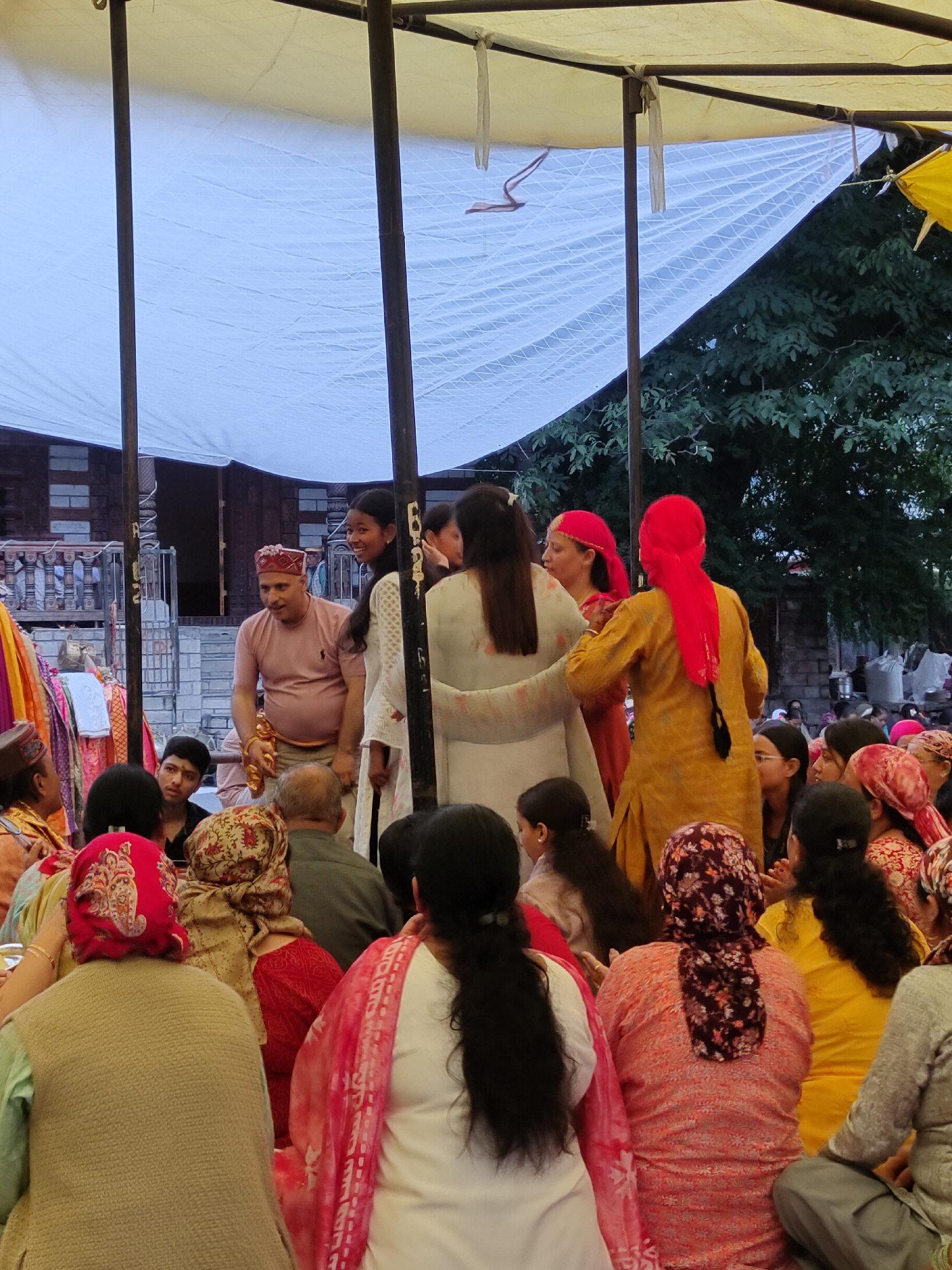 In Siyal, India, a group of people are gathered under a canopy. The canopy is supported by dark poles and is made of a white translucent material. Several individuals are seated in the foreground, facing away from the camera. They are wearing colorful clothing and headscarves. In the middle ground, a man in a pink shirt and a traditional red and gold hat is speaking to a woman in a white dress. Behind them, other people are visible, some also wearing headscarves. The background shows a building with a stone facade and a balcony, along with lush green trees. The overall scene appears to be an outdoor gathering or event.