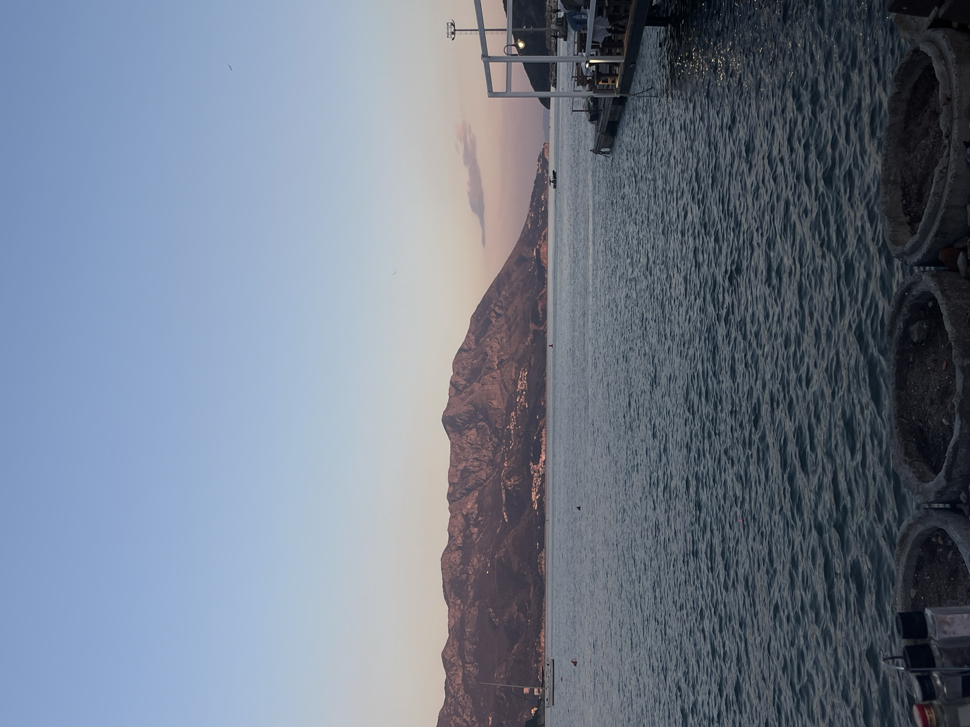 The image captures a scene in Budva, Montenegro, featuring a coastline with mountains rising along the edge of the water. The sky transitions from a pale blue to a soft pink hue, suggesting either sunrise or sunset. The water surface is textured with small waves, and a portion of a dock or pier extends into the water from the right edge of the frame. There are what appears to be concrete structures, possibly part of the waterfront infrastructure, at the far right edge. Along the mountainside, small clusters of buildings or structures are visible. A few small boats are visible on the water in the middle of the image. A small bird is visible in the sky on the left side of the image.