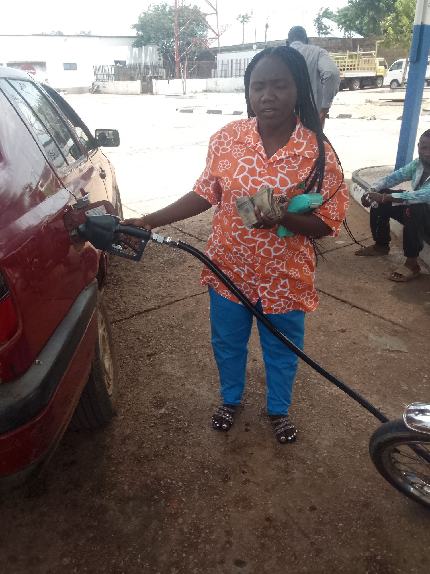 A woman in an orange and white patterned shirt and blue pants stands at a gas station, holding a gas pump nozzle inserted into a red car's fuel tank. In her other hand, she holds a stack of bills and a small green object. She is looking towards the viewer, her expression neutral. Another person is partially visible in the background, and a man is sitting on a ledge or concrete barrier in the background right. A motorcycle is on the right of the frame. The setting appears to be Bukuru, Nigeria, based on the style of buildings and vehicles present in the background. The environment is a paved gas station lot. A truck is visible in the background.