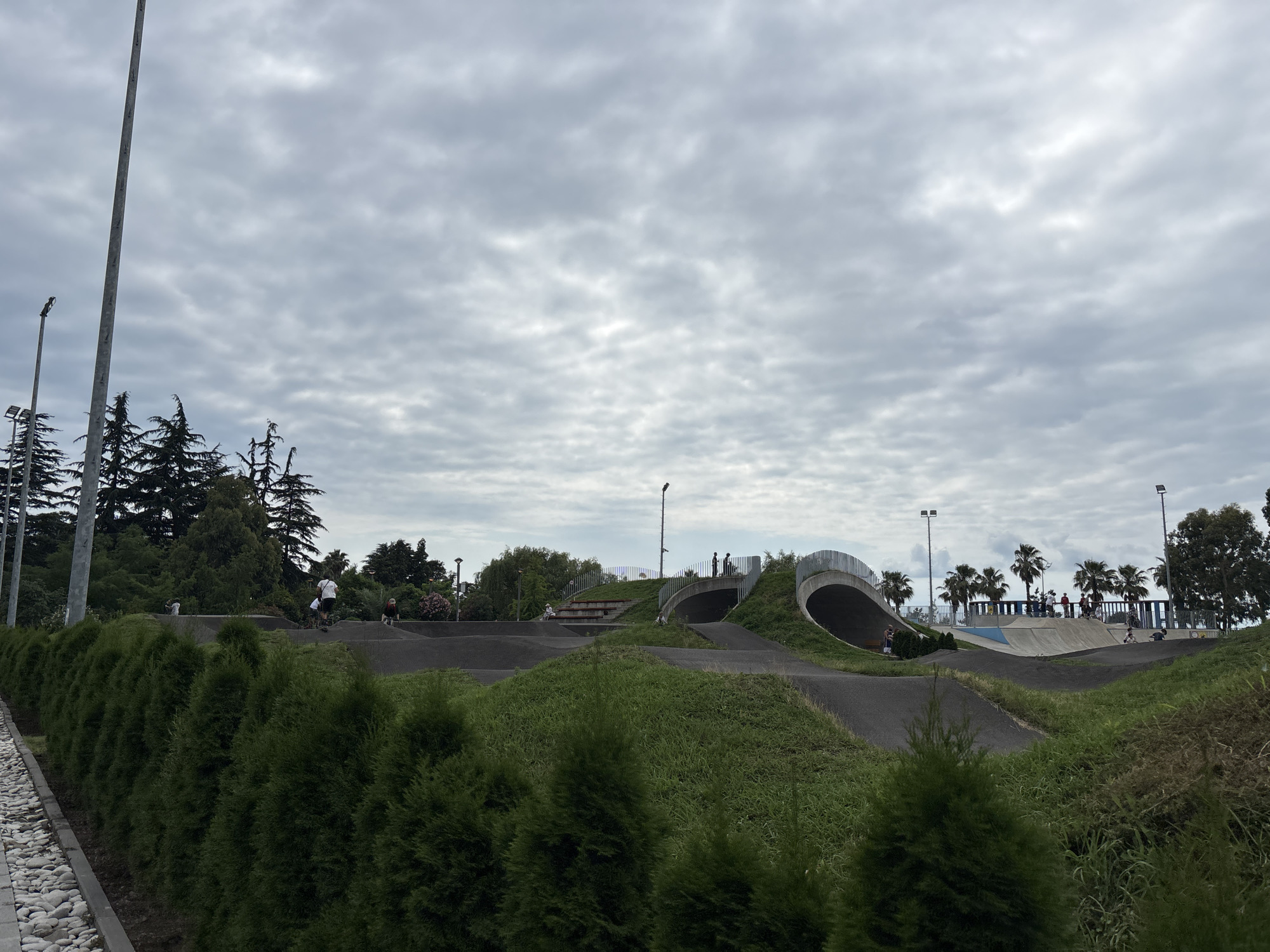 This image depicts a BMX track located in Batumi, Georgia. The track features undulating terrain with asphalt surfaces, surrounded by lush green grass and cypress trees. In the background, there is a concrete structure resembling a tunnel or bridge, and further beyond, palm trees and a skate park area are visible. Several people are present on the track, some of whom appear to be cycling or skateboarding. The sky is overcast with thick clouds. A tall lamppost stands on the left side of the frame, and another in the distance. The foreground is dominated by a row of evergreen shrubs, with a cobblestone path to the left of them.