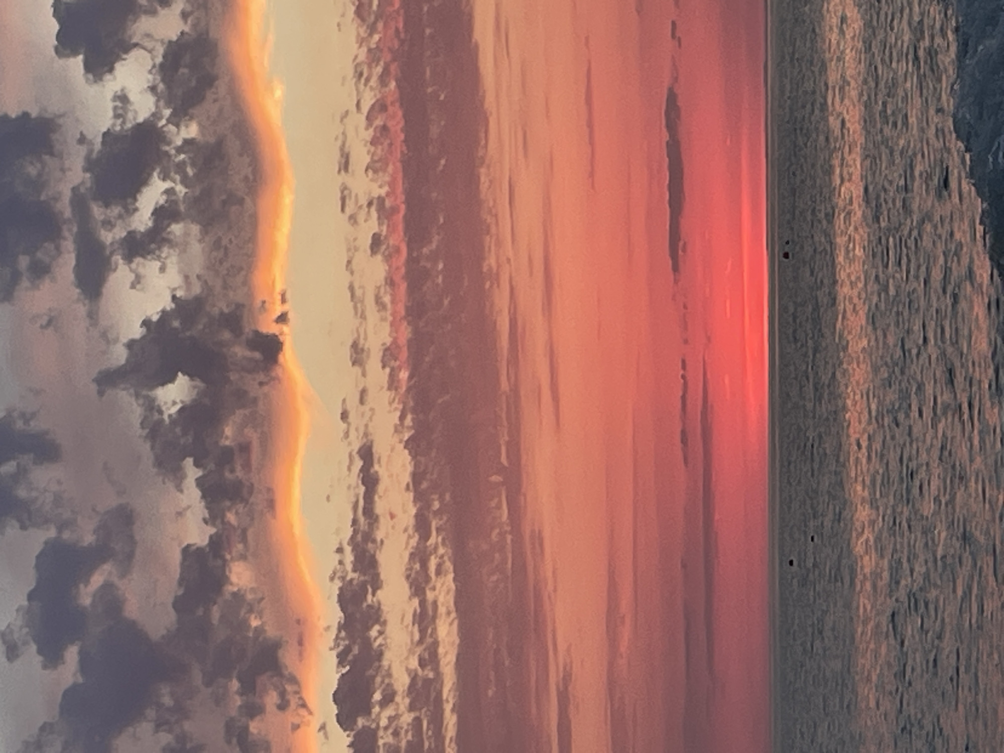 This media file captures a serene seascape at sunset. The scene is dominated by a dramatic sky filled with clouds in varying shades of orange, pink, and gray, indicative of the golden hour. A bright, luminous band of orange-yellow light, likely the setting sun, forms a vivid boundary between the clouds and the water.
The water itself is a textured expanse of gray and blue, reflecting the subdued light of the sky and creating a rippling effect. A distinct horizon line divides the sky and sea. On the right side of the image, near the horizon, there are two small, dark figures that might be people or small objects, though their details are indistinct.
The overall mood is tranquil and picturesque, emphasizing the beauty of nature's twilight display. The time of day is clearly sunset, and the weather appears calm with the presence of clouds. No text is visible in the image.