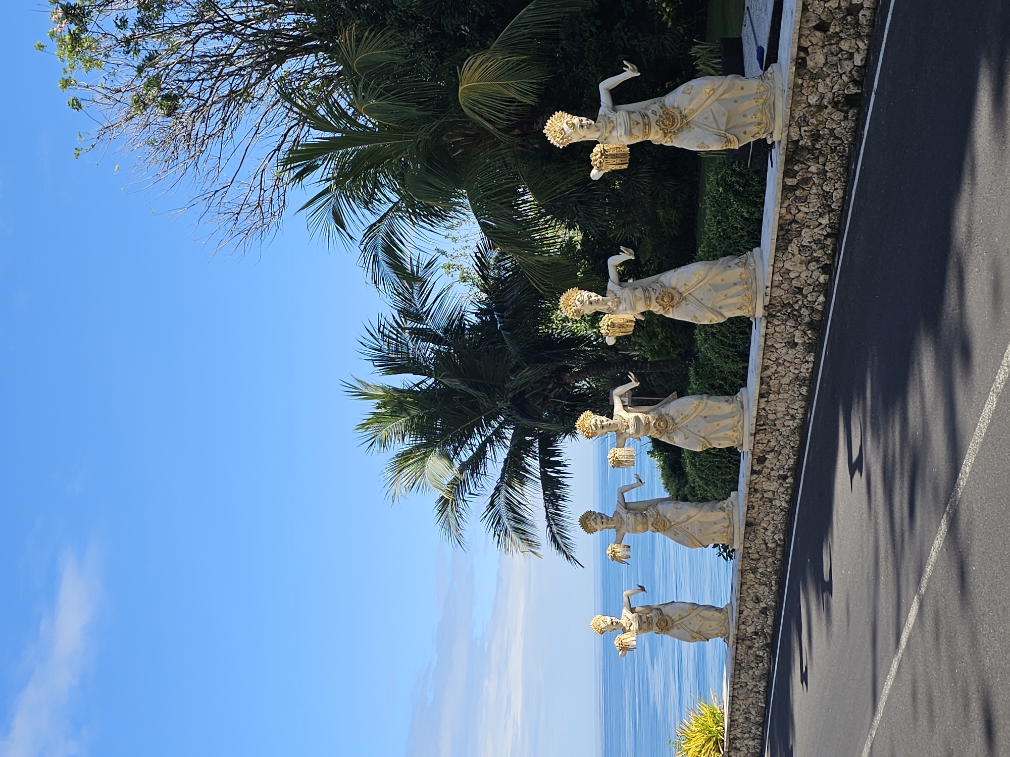 The image presents a roadside scene in Kuta Selatan, Indonesia. There are five stone statues of Balinese dancers lined up on a low stone wall separating the road from the shoreline. The statues are light in color with golden accents. Tropical foliage, including palm trees, provides a backdrop. Adjacent to the wall is an asphalt road with painted lane markers. Beyond the statues, there is a clear view of the ocean under a partly cloudy, blue sky. A bare-branched tree occupies the upper left corner of the frame.
