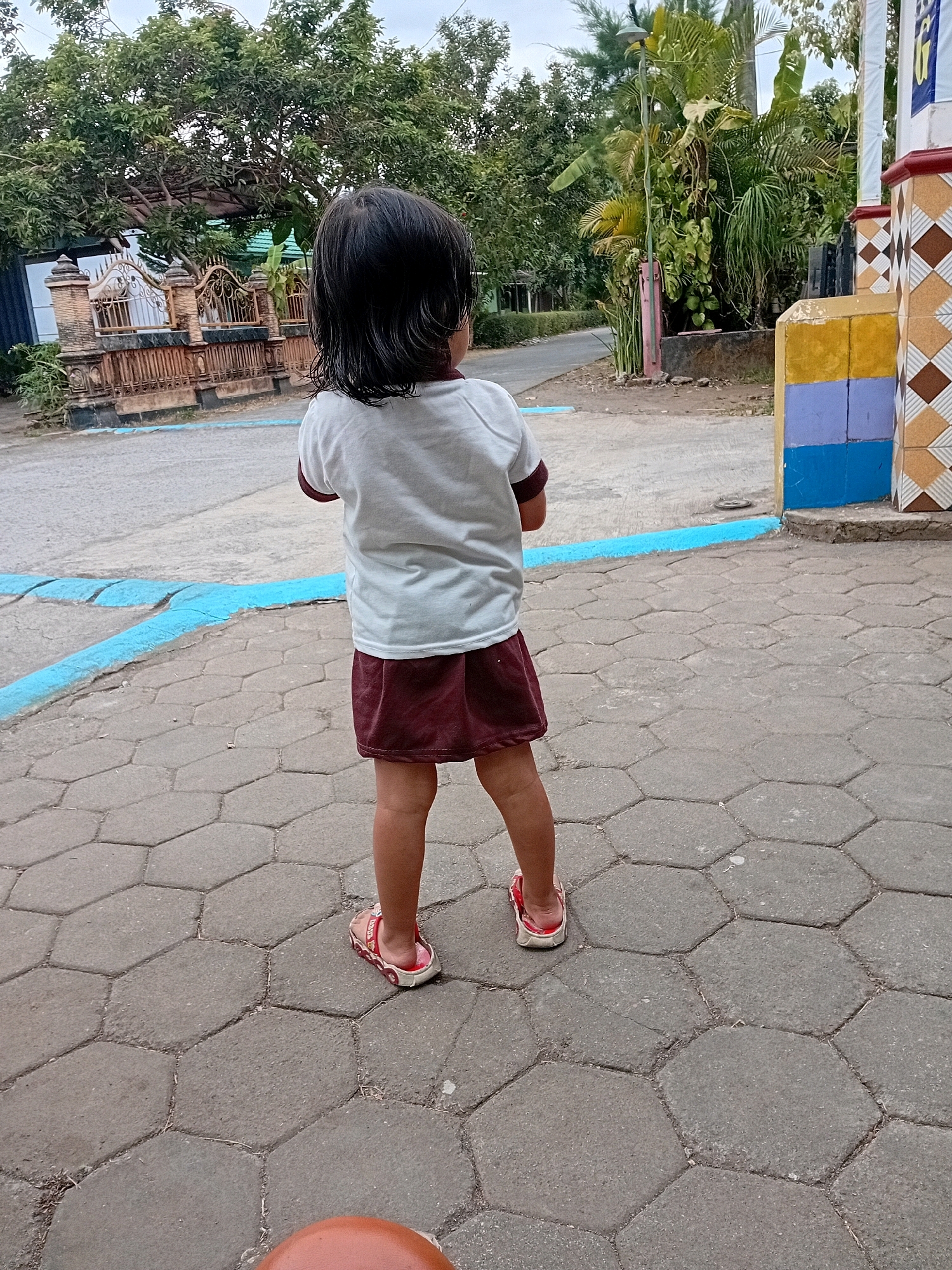 A full shot shows a young child from behind, standing on a hexagonal paved walkway. The child has dark hair and is wearing a white t-shirt with a burgundy collar and a matching burgundy skirt. They are also wearing red sandals. The walkway is bordered by a blue painted line. In the background, there is a street, a fence, and lush green trees. To the right of the frame, a colorful building facade with yellow, blue, and brown tiles is visible. The scene is likely located in Gebang, Indonesia, as indicated by the environment and architectural elements.