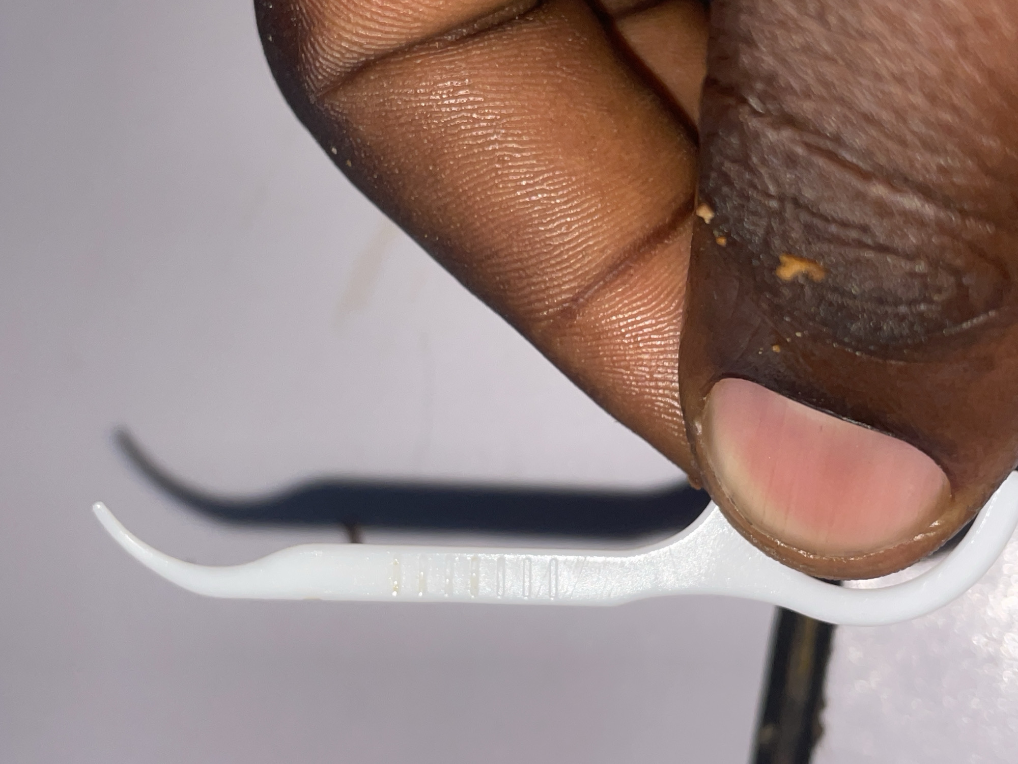 The image shows a close-up view of a person's hand holding a white dental floss pick. The pick has a curved tip and a textured grip. The hand is dark-skinned and appears to have some food particles on the fingers. The background is a plain, light-colored surface. There are no discernible landmarks or details that would indicate a specific location in Jos, Nigeria. The image is purely a close-up of the hand and the dental floss pick.