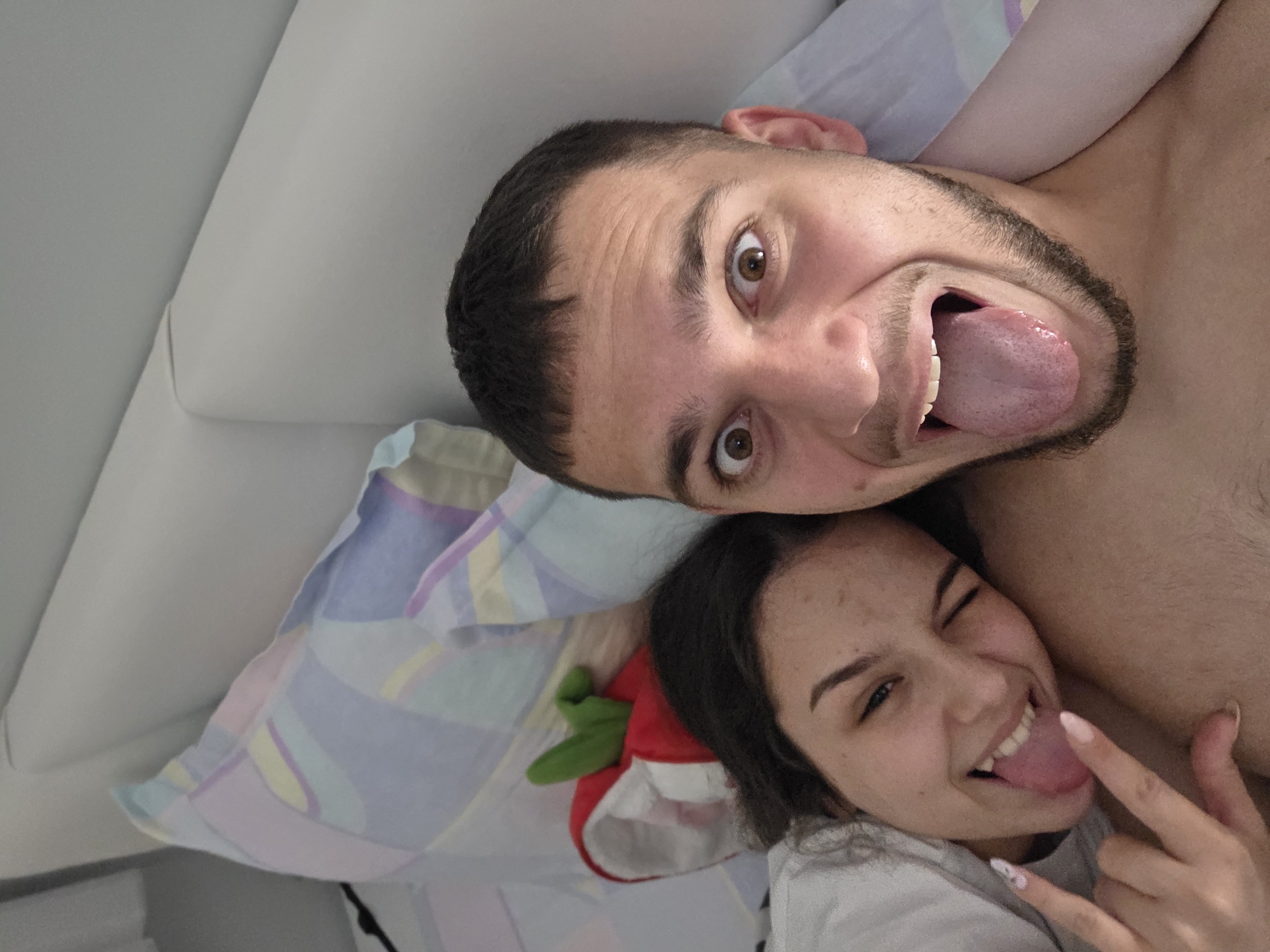 The image displays two young adults, a male and a female, captured in a close-up, likely self-portrait, within an indoor bedroom setting, consistent with a residence in Stara Zagora, Bulgaria. The male, positioned centrally, has short dark hair and light facial stubble. His eyes are wide open, and his mouth is agape with his tongue fully extended forward, indicating an exaggerated, playful expression. The female is situated to the left, partially obscured and positioned closely to the male. She has dark hair, is winking with her right eye, and is smiling broadly with her tongue also extended. Her left hand is raised, forming a "rock-on" gesture. She wears a light-colored garment. Behind the individuals, a pillow with a pastel multi-colored geometric pattern (blues, purples, yellows, pinks) is visible. To the upper left, behind the female's head, a red object with a green stem-like top, possibly a plush toy, is partially visible. A plain white headboard or wall forms the background.