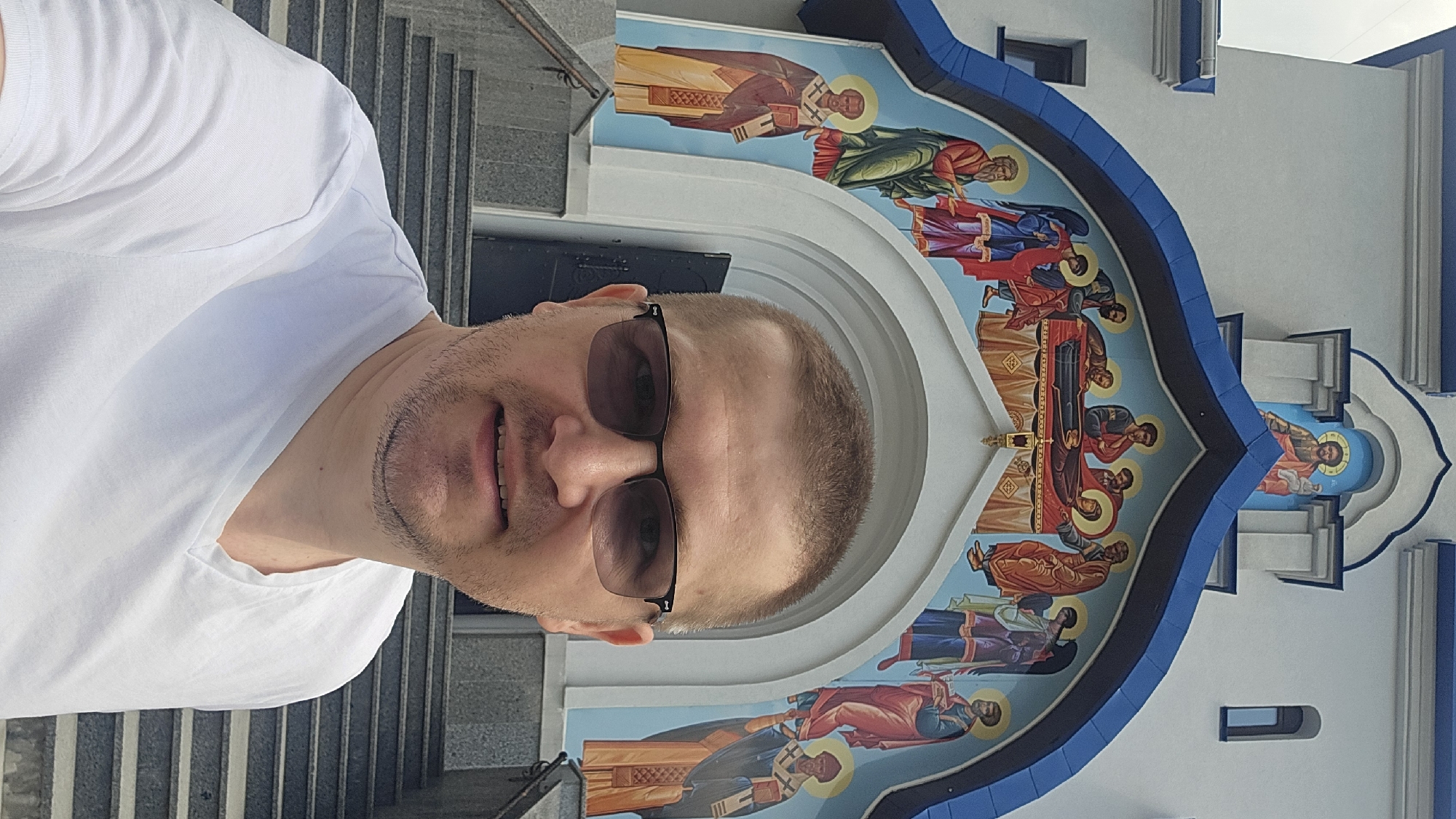 A man is taking a selfie in front of a church. The church has a large fresco above the entrance depicting religious figures. The man is wearing a white t-shirt and sunglasses. He has short hair and stubble. The church appears to be Orthodox Christian, given the iconography. The location is the Church of the Archangel Michael in Kyiv, Ukraine.