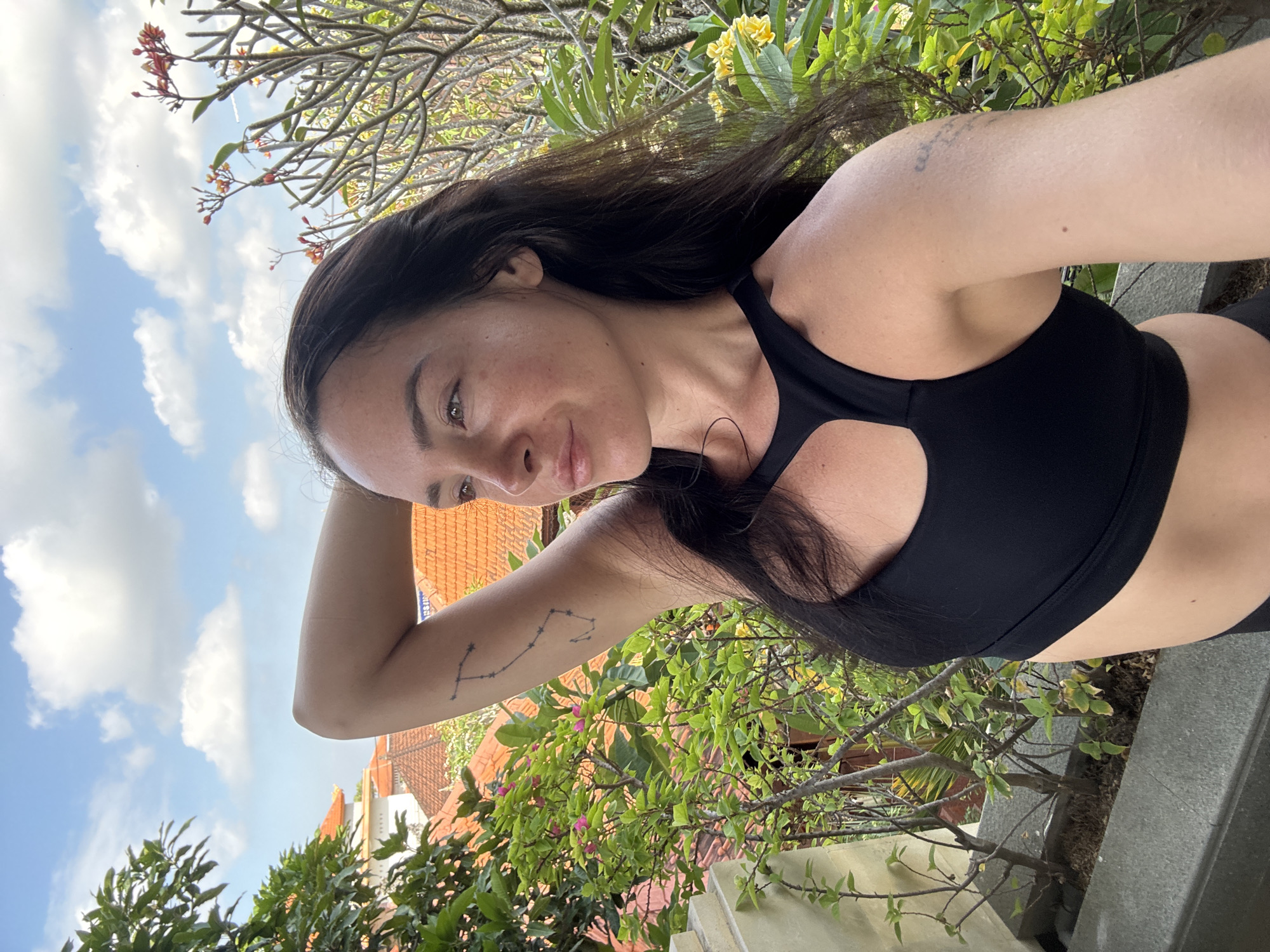 The image shows a woman with long, dark hair taking a selfie outdoors. She is wearing a black sports bra and her left arm is raised, with her hand behind her head. She has a constellation tattoo on her upper left arm and another tattoo on her right arm. The background features lush green foliage, blooming flowers, and tiled rooftops, suggesting a tropical or sub-tropical urban environment. The sky above is blue with scattered white clouds. The composition and surroundings are consistent with a location in North Kuta, Indonesia, known for its vibrant greenery and distinctive architecture.
