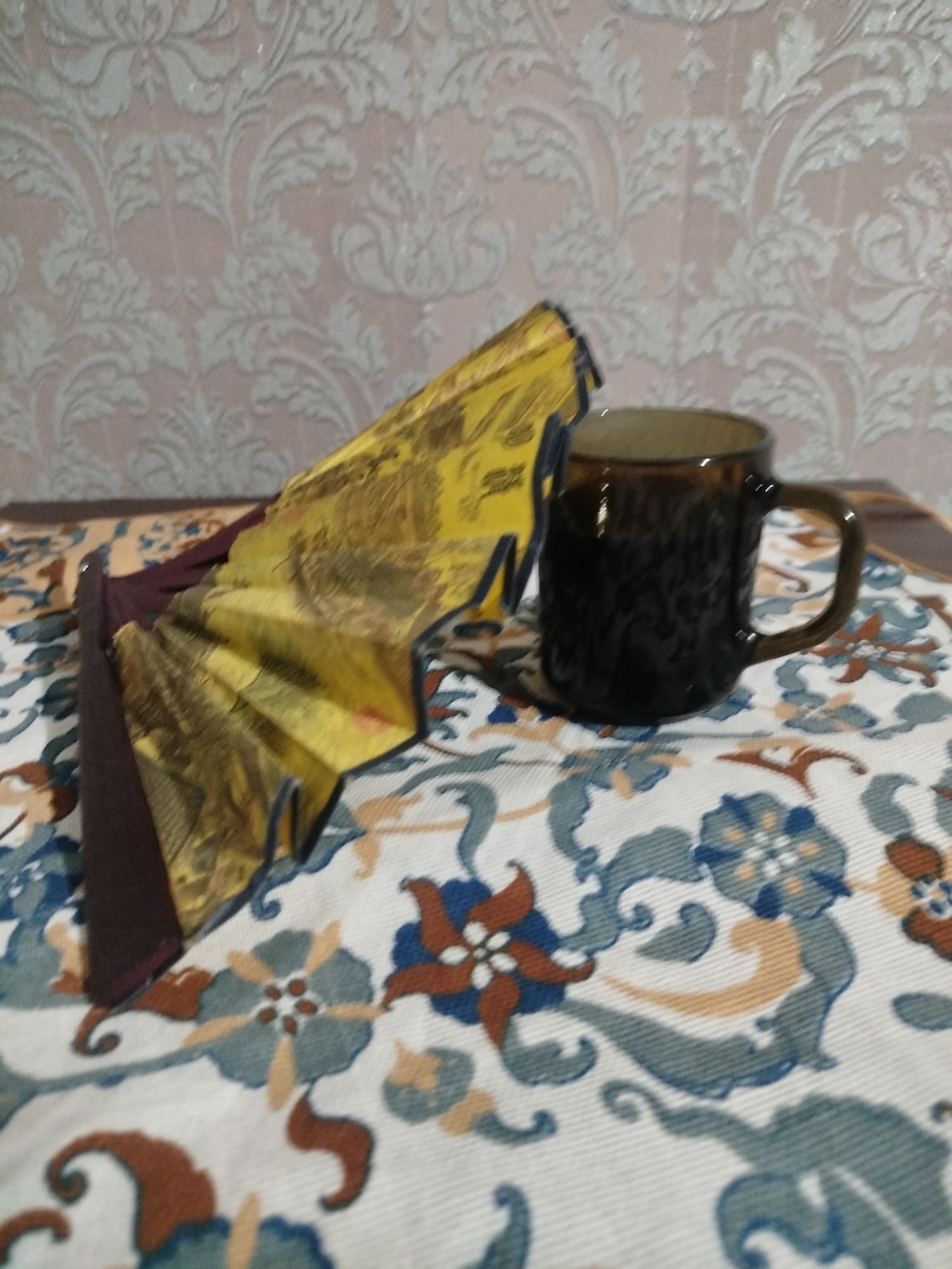 The image shows a close-up of a folded yellow fan with dark brown accents lying on a patterned tablecloth. Next to the fan is a dark brown mug with a handle. The tablecloth has a busy pattern of blue, brown, and cream floral and scroll motifs. In the background, there is a textured wallpaper with a repeating damask pattern in light grey and pinkish hues. The scene is indoors, likely on a table. There are no people or animals present.