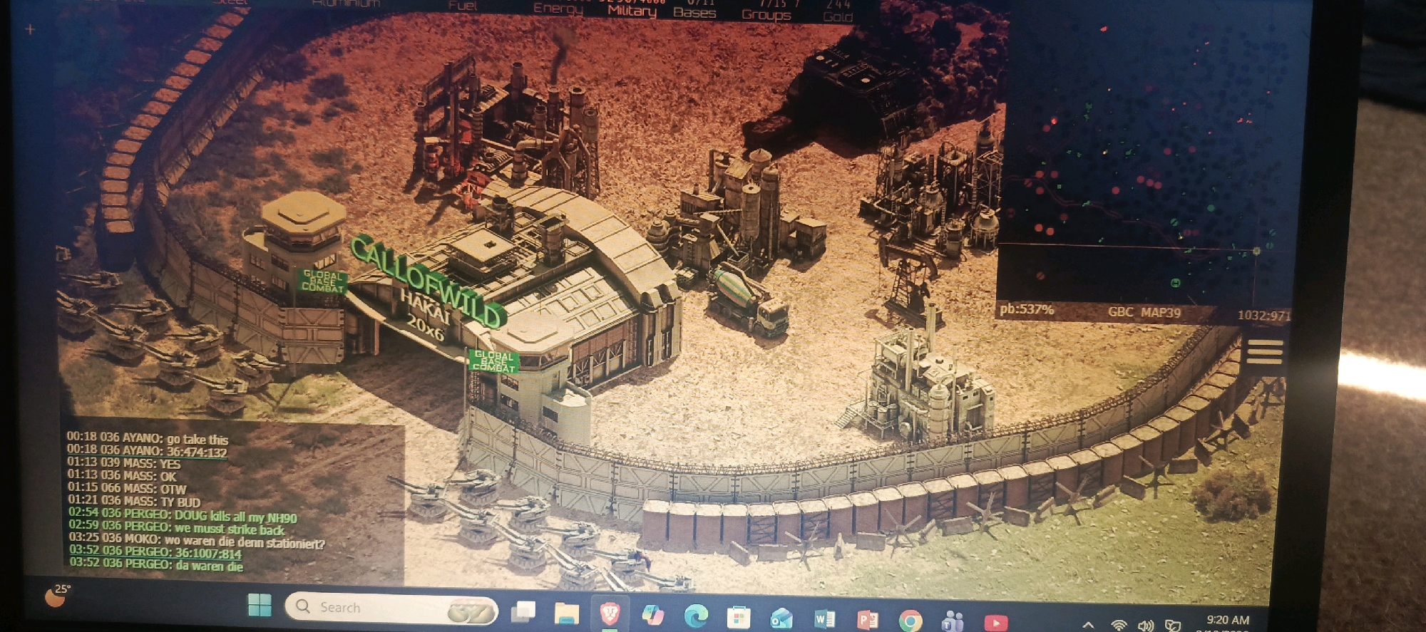 This image depicts a screenshot of a real-time strategy video game, likely set in a desert environment. The foreground shows a fortified military base with a curved wall, watchtowers, and several tanks positioned in front of it. Within the base, various industrial-looking structures, possibly factories or resource processing plants, are visible, along with a cement mixer truck. A prominent sign reads "CALLOFWILD HAKAI 20X6 GLOBAL BASE COMBAT."
A chat log is displayed in the lower-left corner of the screen, with messages exchanged between players using usernames like AYANO, MASS, PERGEO, and MOko. The chat log indicates in-game actions such as "go take this," "we must strike back," and inquiries about unit placement ("wo waren die denn stationiert?").
The upper portion of the screen displays game interface elements, including resource counters (Fuel, Energy, Military, Bases, Groups, Gold) and numerical data like "pb:537% GBC MAP39 1032:971."
The environment is characterized by a sandy, arid landscape with sparse vegetation and an orange-tinted sky, suggesting a desert or post-apocalyptic setting. There is no discernible depiction of Miana Thub, Pakistan, or any real-world locations within the game's visual representation. The scene is purely from a digital simulation.