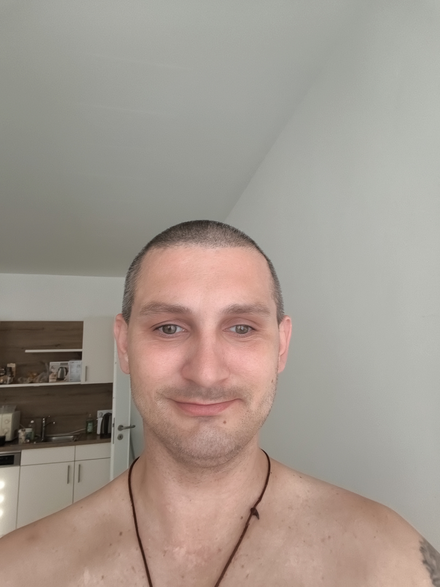 A man with a shaved head is taking a selfie indoors. He is shirtless and wearing a brown cord necklace. His skin has some uneven pigmentation and a scar is visible on his neck. In the background, a kitchen area with white cabinets, a sink, and a stovetop is visible. The walls are painted a light gray and the ceiling is white. The scene is a domestic indoor environment.