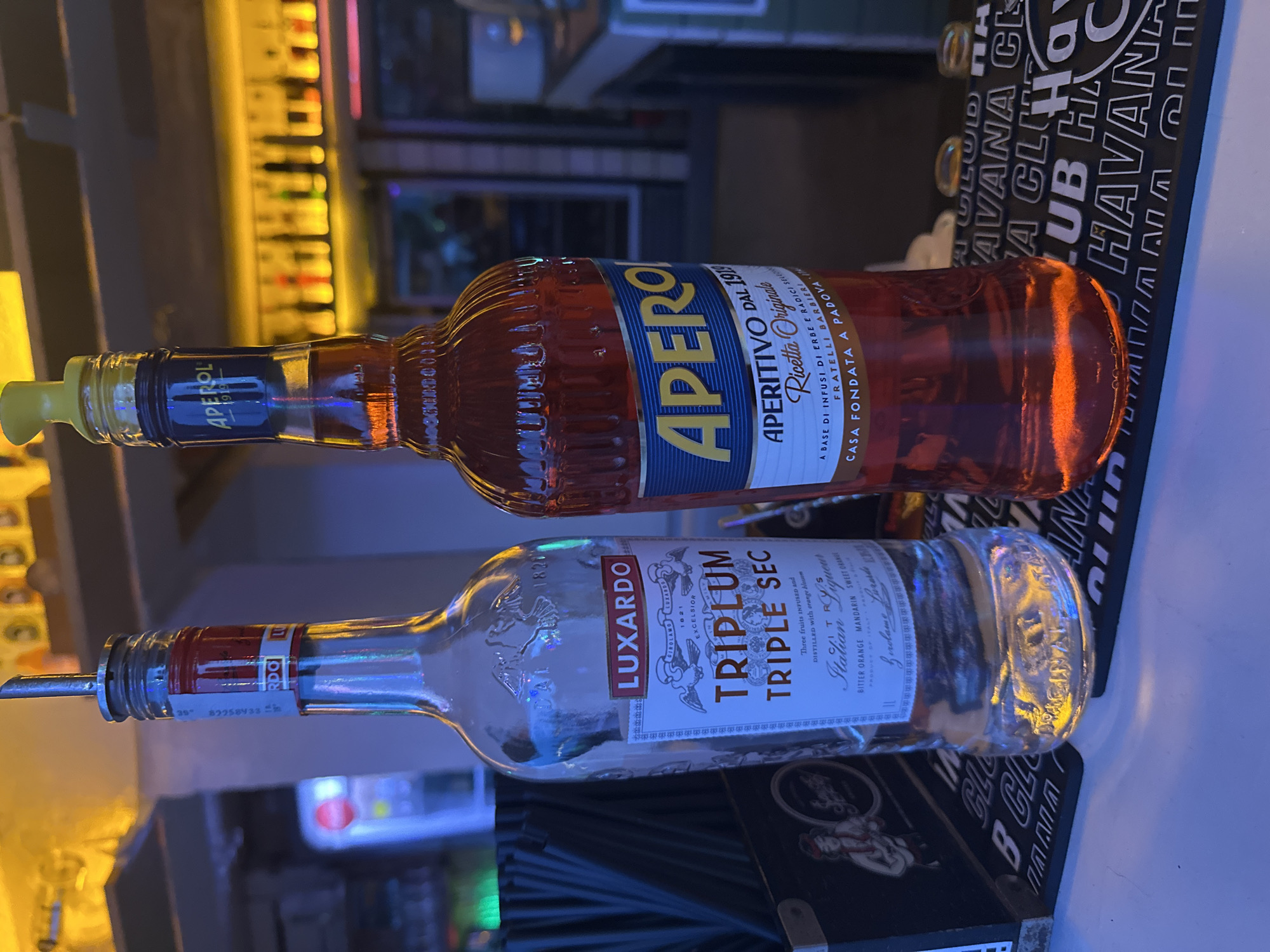 The image shows a close-up, eye-level shot of two bottles of alcohol on a bar counter. The bottle on the left is clear and appears to be Luxardo Triplum Triple Sec, with a red and white label. It has a pour spout inserted into its opening. The bottle on the right is filled with an orange-colored liquid and is identified as Aperol Aperitivo. It has a blue and yellow label and a yellow cap. The background is blurred, with shelves of bottles and colorful lighting, suggesting an indoor bar or nightclub setting. A portion of a black bar mat with the "Havana Club" logo is visible in the foreground. The image is taken in the city of Varna, Bulgaria.