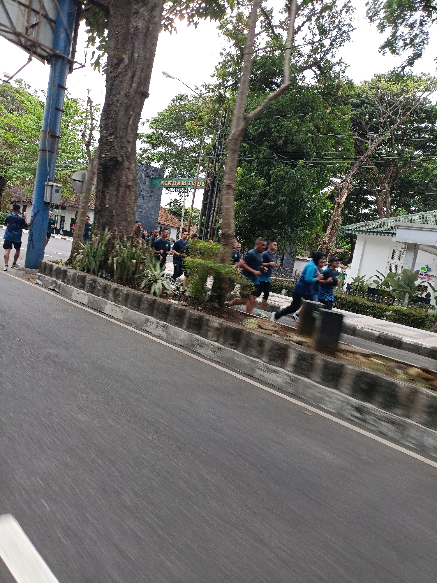 A group of men in matching dark blue athletic wear are running along a paved road in a city. The men are blurred due to motion, but appear to be of various ages and build. They are running in a group, with some in front and others trailing behind. They are running on a sidewalk or shoulder next to the road. Behind them is a line of concrete barriers, vegetation, trees, and buildings. The buildings appear to be a mix of white and brick, with one white building having a green roof. There is a prominent large tree in the foreground on the left side of the image, with a blue pole next to it. The road itself is paved and gray. The scene suggests an outdoor athletic event or training session in an urban environment. The presence of the sign "RINDAM IV/DIPONEGORO" on a dark gray structure in the background indicates the location is within the Rindam IV/Diponegoro Military Academy in Magelang, Indonesia.