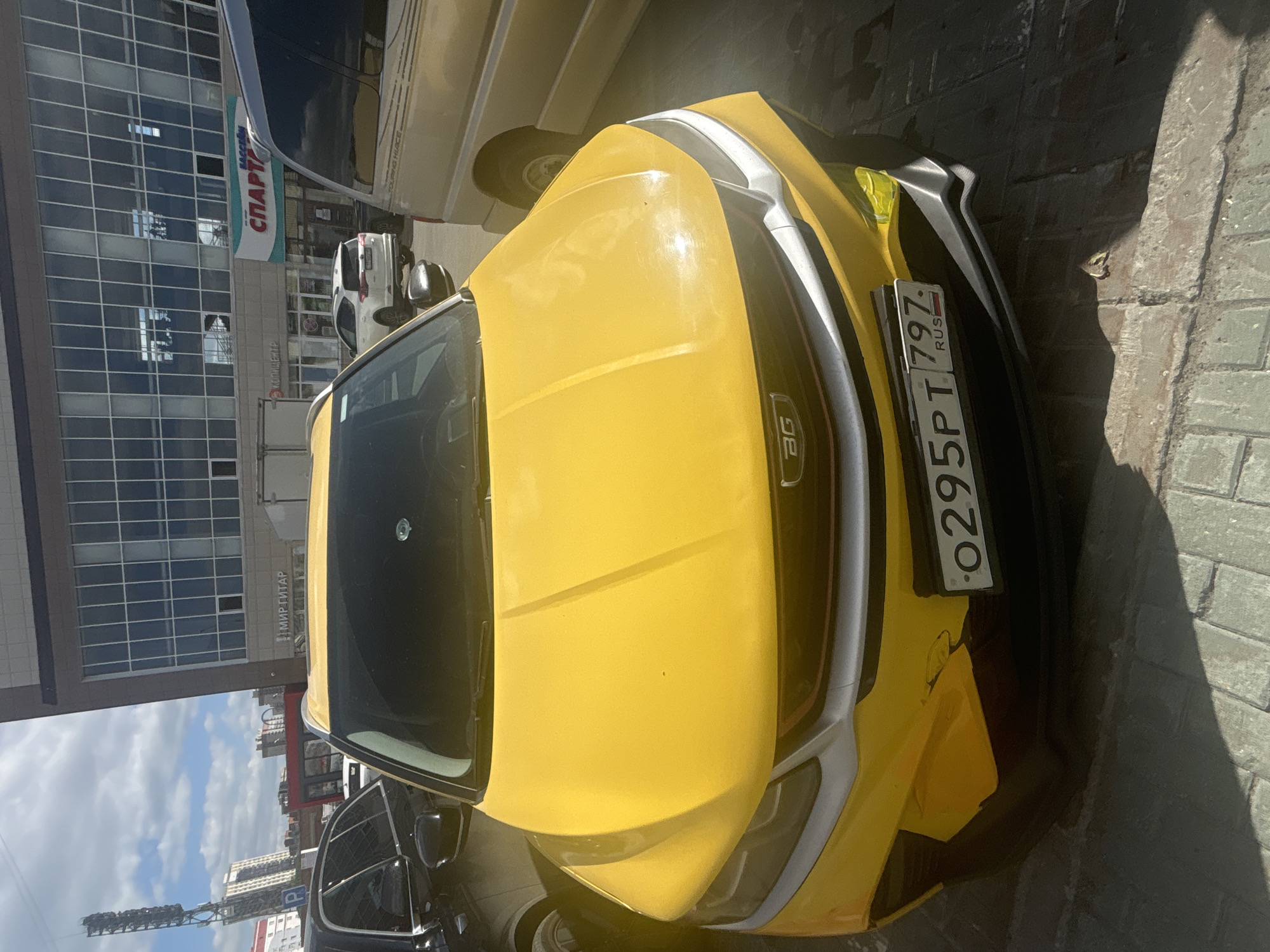 A yellow sedan with a license plate reading "O295PT 797 RUS" is parked on a street in front of a modern building in Novosibirsk, Russia. The building features large glass windows and a sign that reads "СПАРТА" which translates to "Sparta". A black SUV is parked to the left of the yellow sedan, and a tan van is parked to its right. The ground is paved with light-colored bricks. The sky is blue with scattered clouds. The scene appears to be a daytime street view.