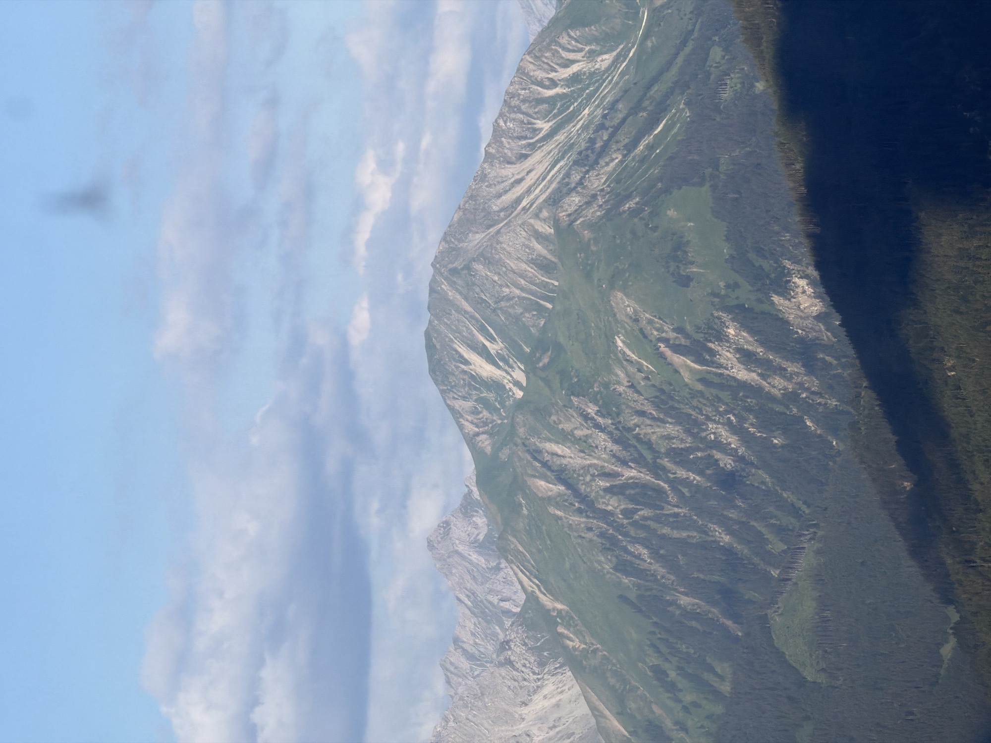 The image shows a wide shot of mountainous terrain under a bright blue sky with scattered white clouds. In the foreground, a densely forested mountain slope is visible, partially covered in shadow. Beyond this, a large mountain with green, grassy slopes and rocky outcrops dominates the mid-ground. The upper slopes of this mountain are streaked with lighter-colored rock. Further in the background, the jagged, rocky peaks of more distant mountains rise, appearing lighter in color and texture. The overall impression is one of a vast, natural landscape. The provided crops show close-ups of the sky with clouds and darker, out-of-focus elements, and detailed views of the mountain slopes, highlighting the textures of rock and vegetation.