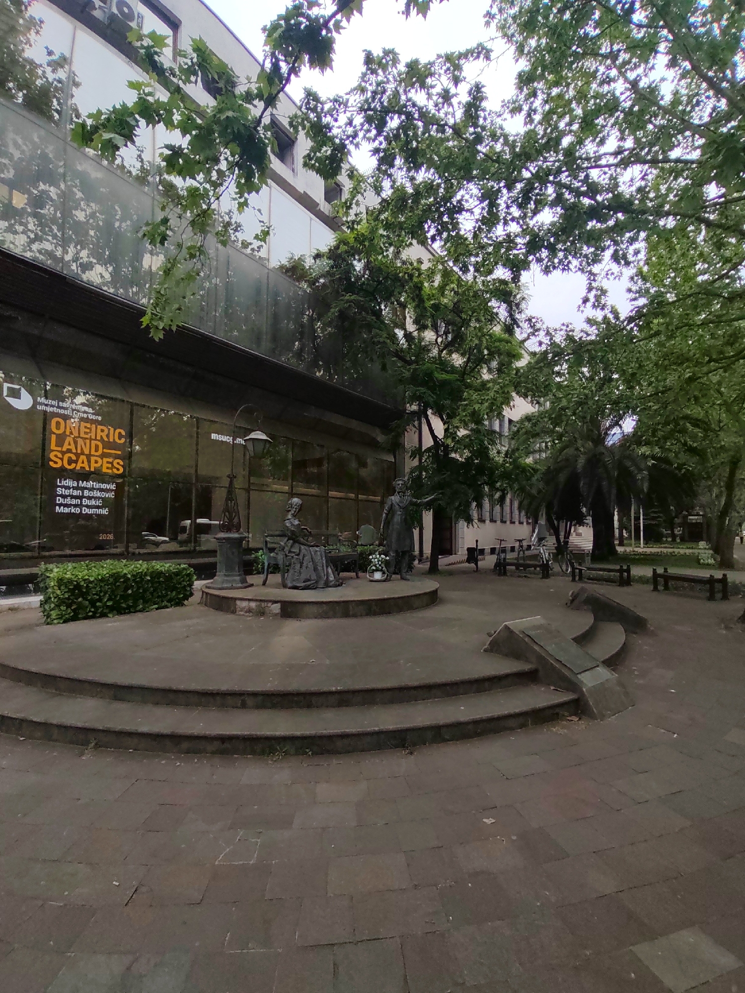 The image shows a public area in Podgorica, Montenegro, featuring a contemporary art museum and several statues. The museum, “Muzej savremena umjetnosti Crne Gore," displays a poster advertising an exhibition titled "ONEIRIC LANDSCAPES," running until 2026. The exhibition features artists Lidija Martinović, Stefan Bošković, Dušan Đukić, and Marko Dumnić.
In the foreground are two statues on circular platforms, accessed by a series of steps. One statue depicts a woman seated on a bench near a lamppost. The other statue shows a man standing with a basket filled with flowers.
In the background, several benches and bicycles are present. Trees are dispersed throughout the area, providing shade. The ground is covered in dark pavement. The overall environment appears to be a public park or gathering space in the city.