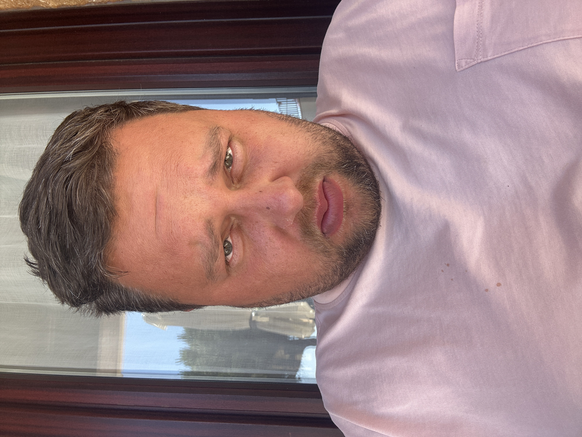 A middle-aged man is pictured from the chest up, wearing a light pink shirt with a pocket. He has short, graying hair, a beard, and fair skin. His eyes are open, and he's looking at the camera. The background appears to be a window with a dark brown frame, and reflections suggest an outdoor scene. It is not possible to know the location in the city of Sant Feliu de Guíxols, Spain.