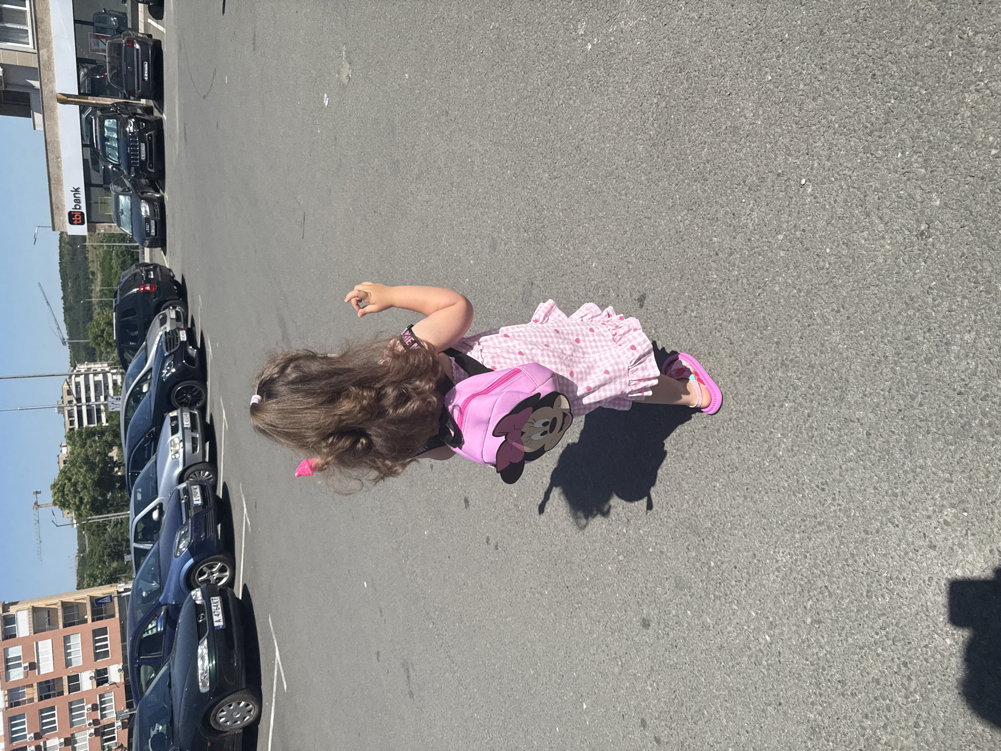 In Burgas, Bulgaria, on a sunny day, a young girl with long, curly brown hair is walking away from the camera in a parking lot. She is wearing a pink checkered dress with ruffles and a pink backpack adorned with a Minnie Mouse design. She has pink sandals on. The parking lot is paved with asphalt and is filled with various cars, including a dark green Opel Vectra, a blue Opel Astra, a silver Opel Astra, a black Audi, a black Jeep Grand Cherokee, and a black Mercedes-Benz. In the background, there are buildings, including a multi-story residential building and a building with the TBI Bank logo. Trees and a construction crane are also visible in the distance. The scene appears to be a typical urban environment on a bright day.