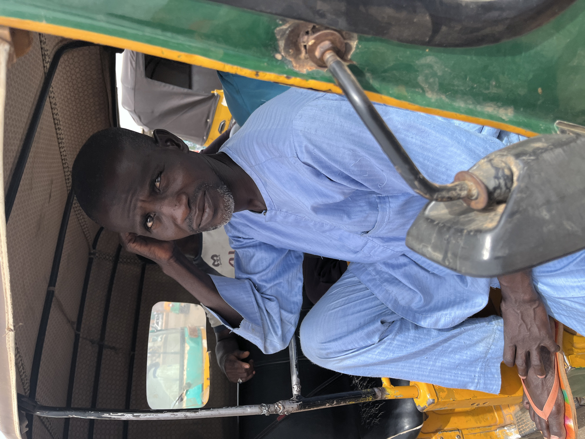 A man with dark skin and a graying beard is seated inside a commercial tricycle in Gura Top, Nigeria. He wears a light blue, long-sleeved shirt and has a pair of orange and white flip-flops on his feet. He is leaning his head on his hand, looking toward the camera. A side mirror of the tricycle is visible in the foreground, and the interior of the vehicle is a beige color. A glimpse of another person can be seen in the background. The tricycle has a green and yellow exterior.