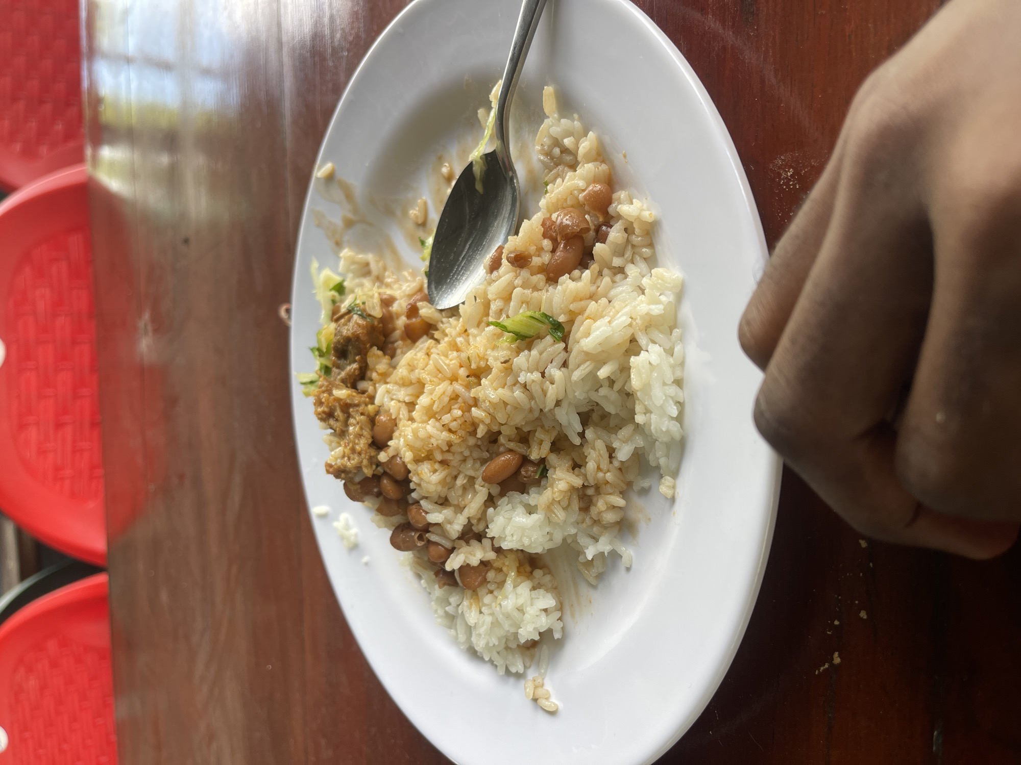 A close-up, high-angle photograph captures a partially eaten meal served on a white, slightly curved plate, resting on a glossy, reddish-brown wooden table. The focus is on the food, with elements of the surroundings and a person's hand visible.
The main dish consists of white rice mixed with light brown beans and a brownish, saucy protein component, possibly a stew or ground meat, along with small pieces of green vegetables or herbs. A silver metal spoon is nestled in the center of the plate, indicating the meal is being consumed. A few grains of rice are scattered on the plate and on the wooden table, suggesting an active eating process.
To the left of the plate, in the background, segments of two red plastic chairs or stools with a patterned surface are visible, hinting at a casual dining environment. The glossy surface of the wooden table reflects ambient light, suggesting it is daytime. On the right side of the frame, a person's dark-skinned hand is partially visible, reaching towards the plate, further confirming the act of eating.
The setting appears to be an indoor or shaded outdoor space, likely a local eatery or a home in Dar es-Salaam, Tanzania, consistent with the type of meal presented. No specific emotions are discernible, and no text is visible in the image.
