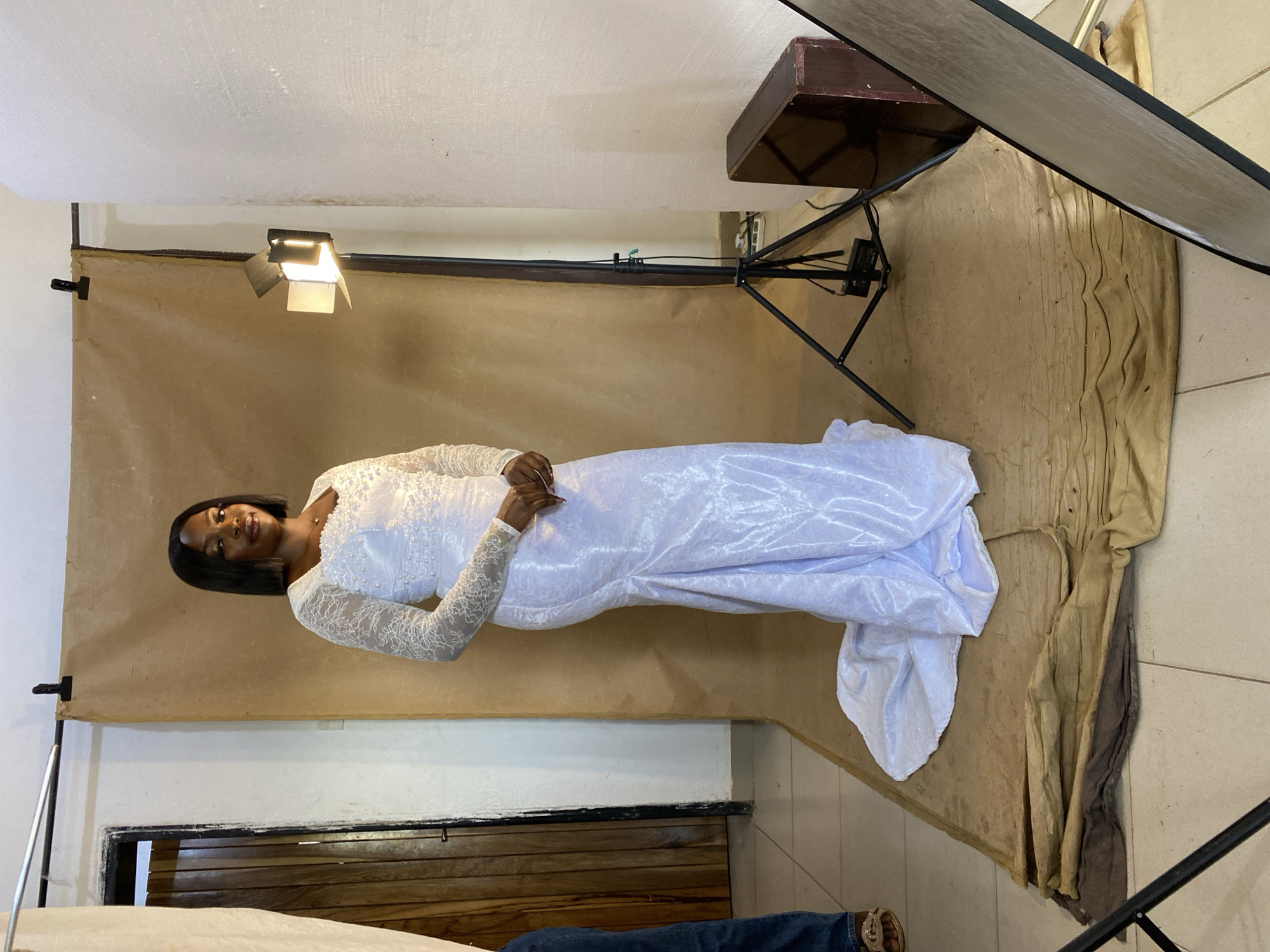 A woman in a long, white, lace-sleeved dress lies on a backdrop of brown and tan fabric. The backdrop is set up in what appears to be an indoor photography studio in Bukuru, Nigeria. A light is positioned above the backdrop on the left side of the frame. A dark-colored box is perched atop a black tripod near the upper right corner. A photography reflector stands on the right side of the frame. A glimpse of blue jeans and tan sandals can be seen in the bottom center.